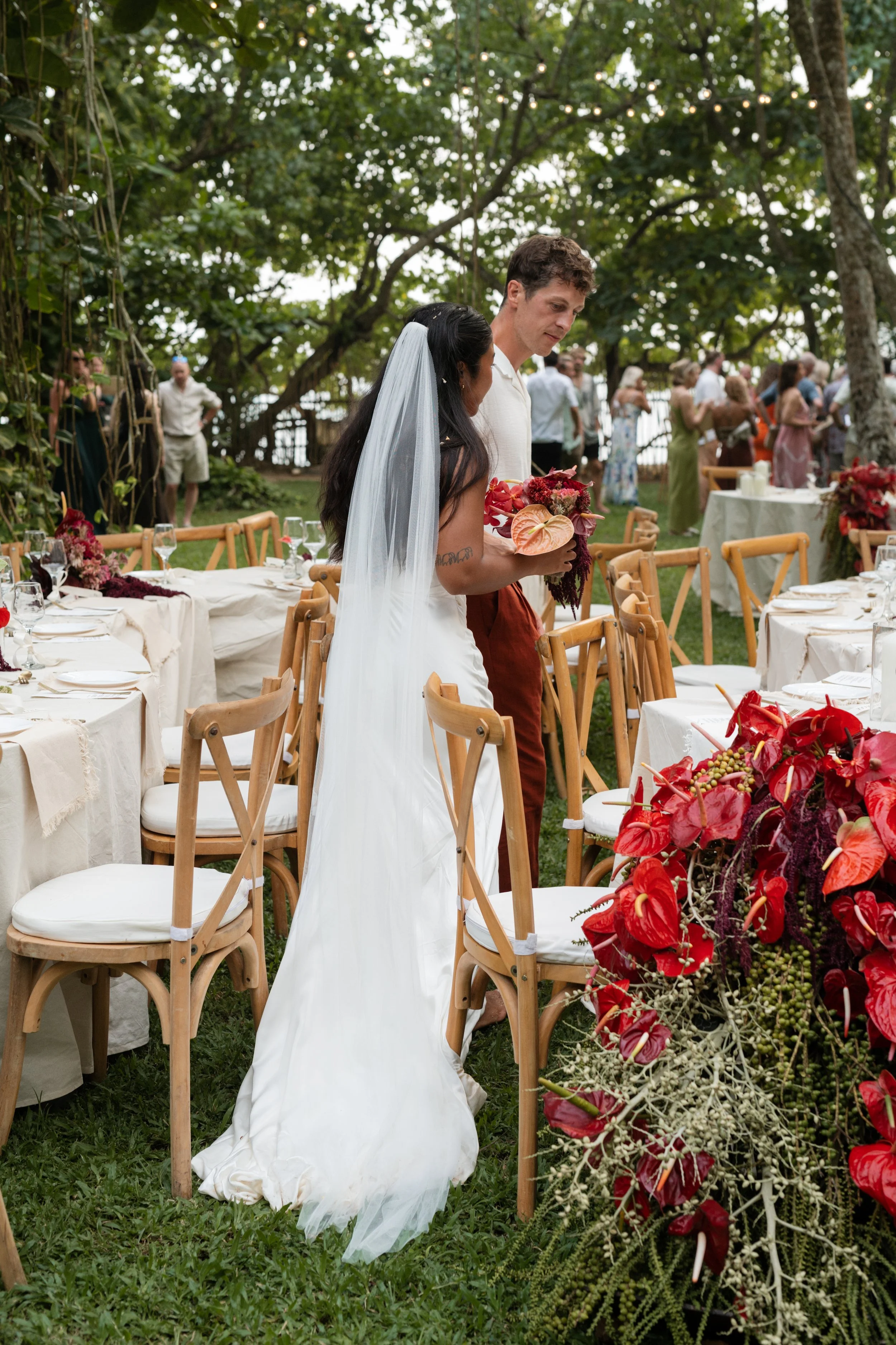 A bride and groom standing together at a wedding reception outdoors, with the bride holding a bouquet of flowers and the groom looking down. The scene includes decorated tables and chairs, with other guests in the background, surrounded by trees.