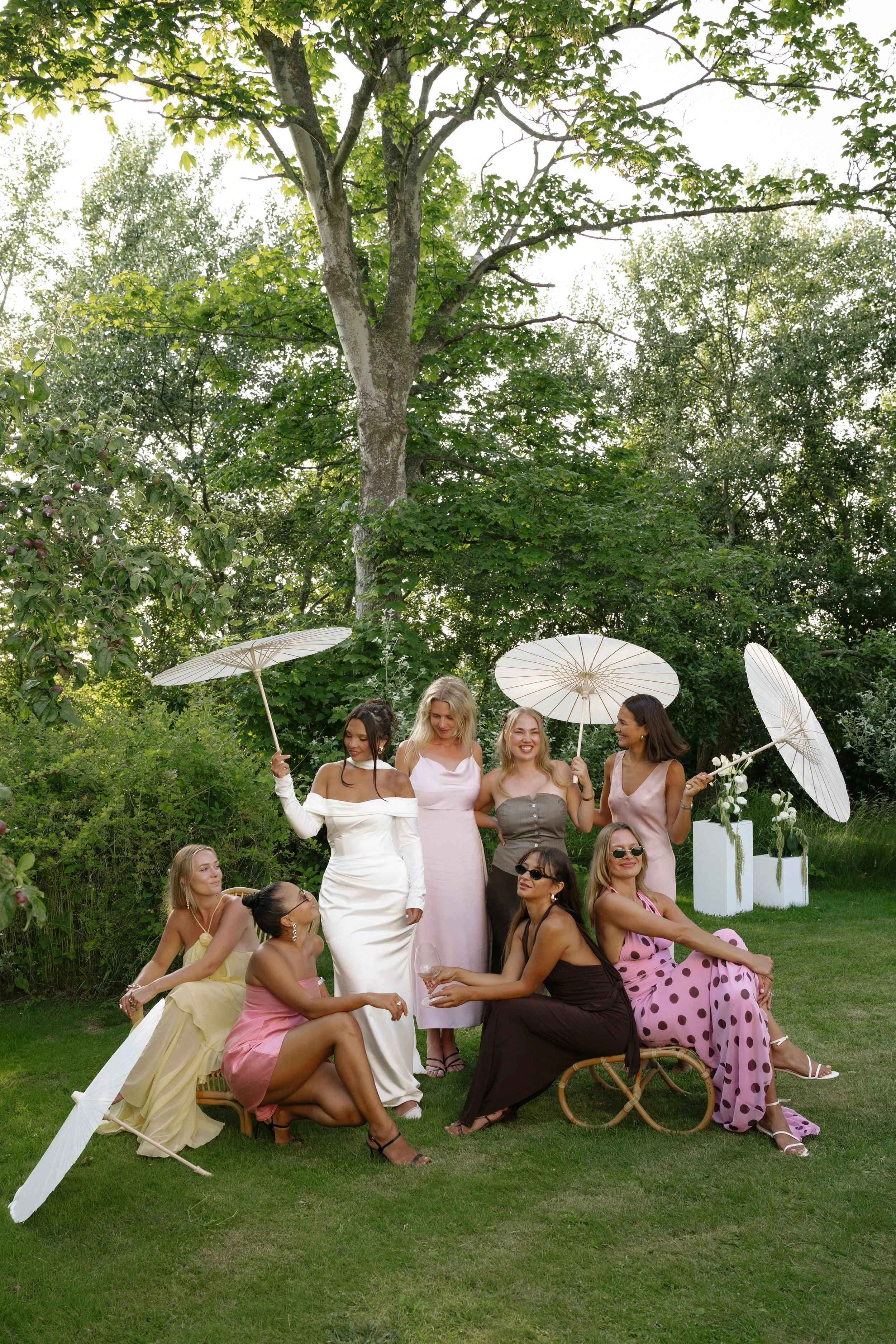 A group of eight women enjoying a garden party, some sitting and some standing, holding white parasols, dressed in colorful and elegant summer dresses, with greenery and trees in the background.