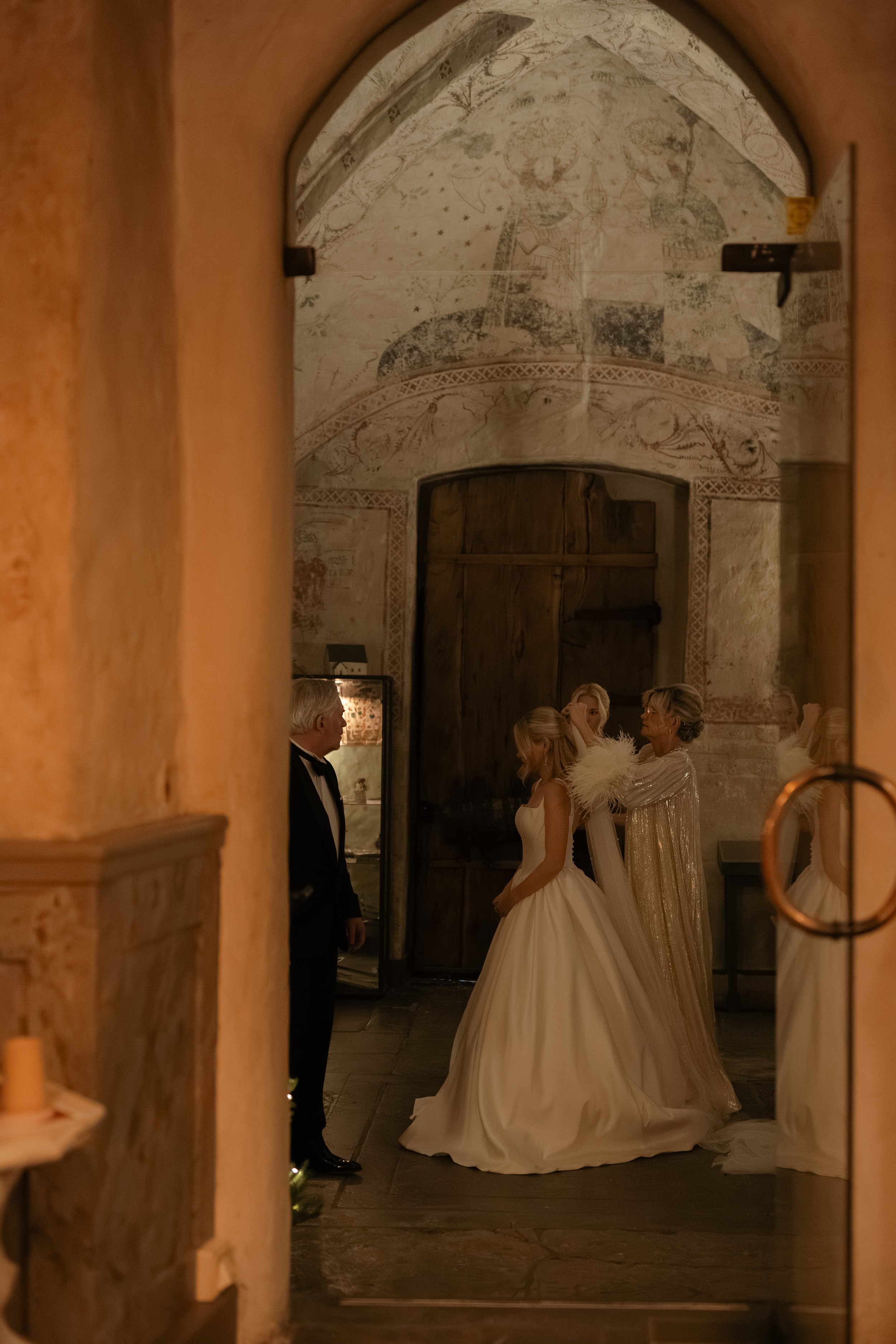 A wedding scene seen through a doorway, with a bride in a white gown, two women, and a man in a tuxedo inside a rustic, historic building with detailed wall illustrations.