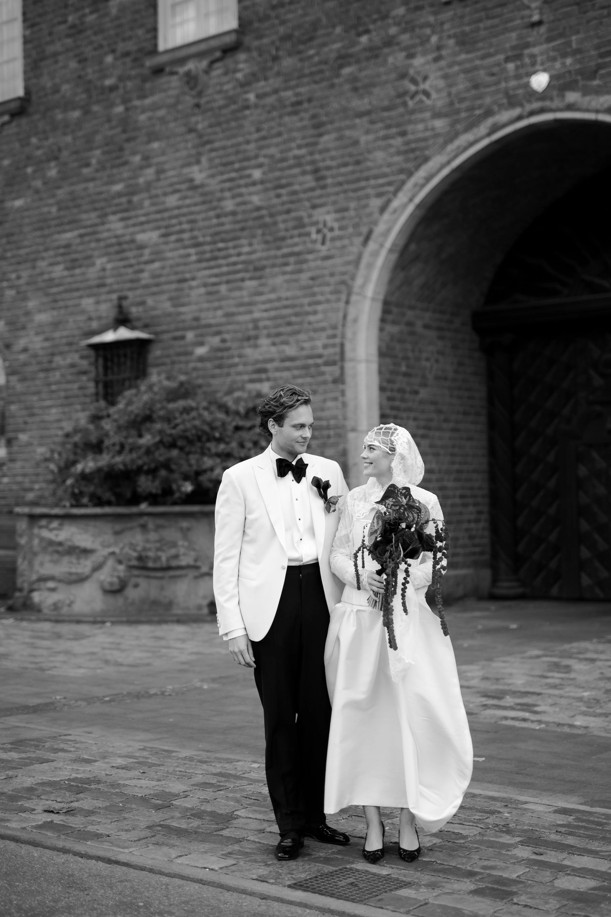 Black and white photo of a bride and groom walking together outside near a brick archway, with the groom in a tuxedo and the bride in a vintage white wedding dress and lace head covering, holding a large bouquet of flowers.