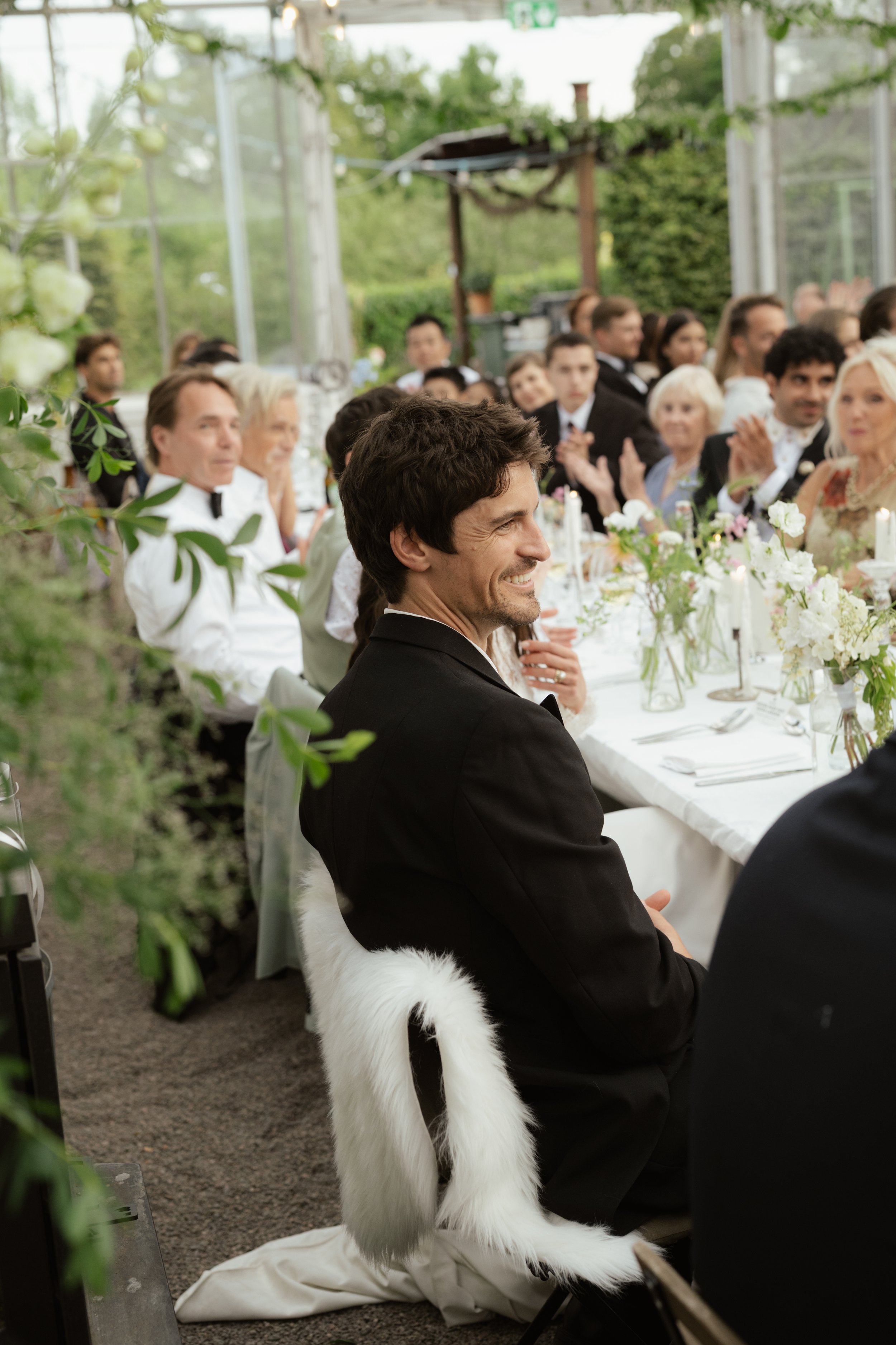 A man seated at a formal event, smiling and dressed in a black tuxedo, with a furry white and black animal tail attached to his chair. The event is held in a bright, glass-walled venue with hosted guests seated at long tables decorated with flowers.