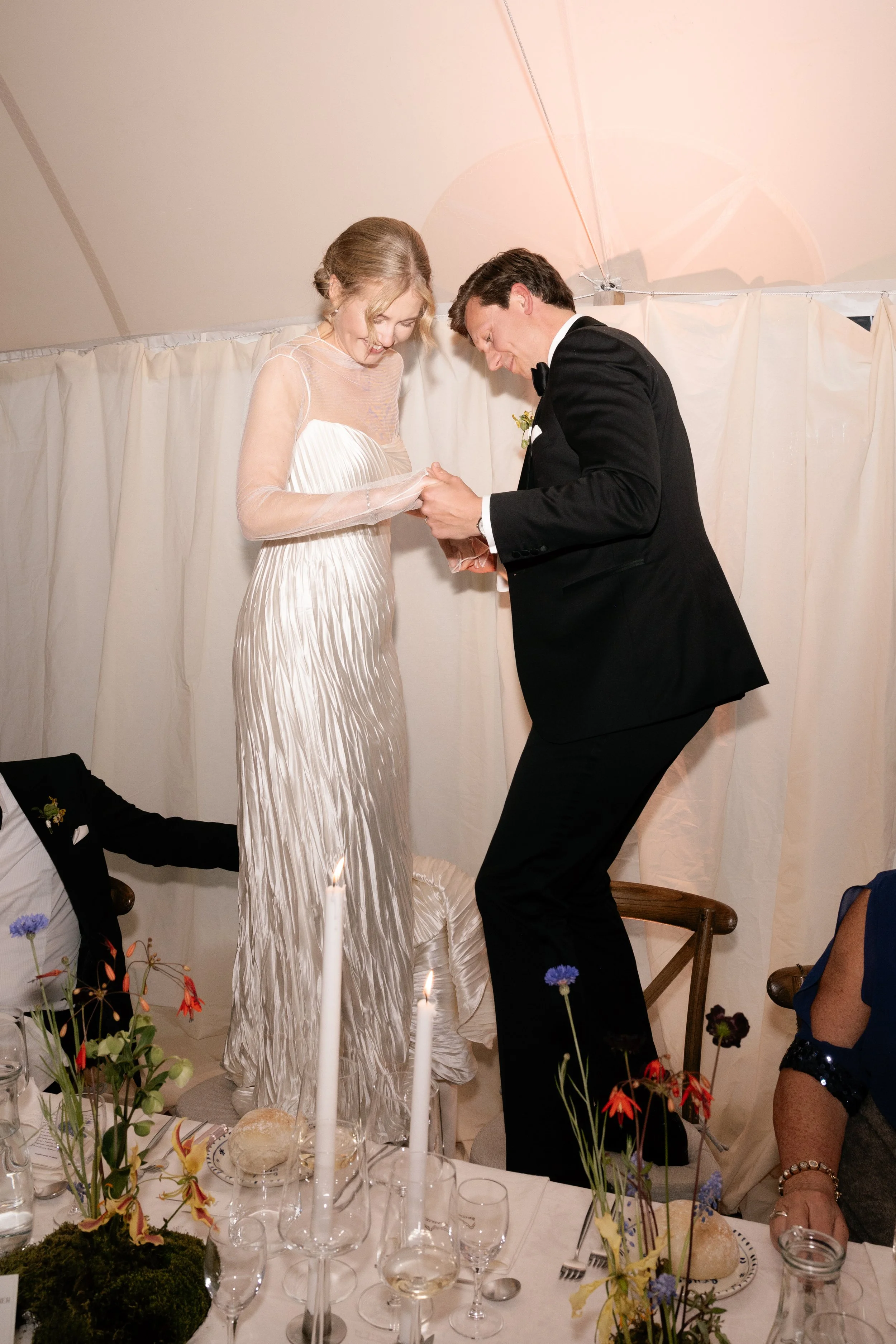 A bride and groom on a table, holding hands, stand on chairs during their wedding celebration.