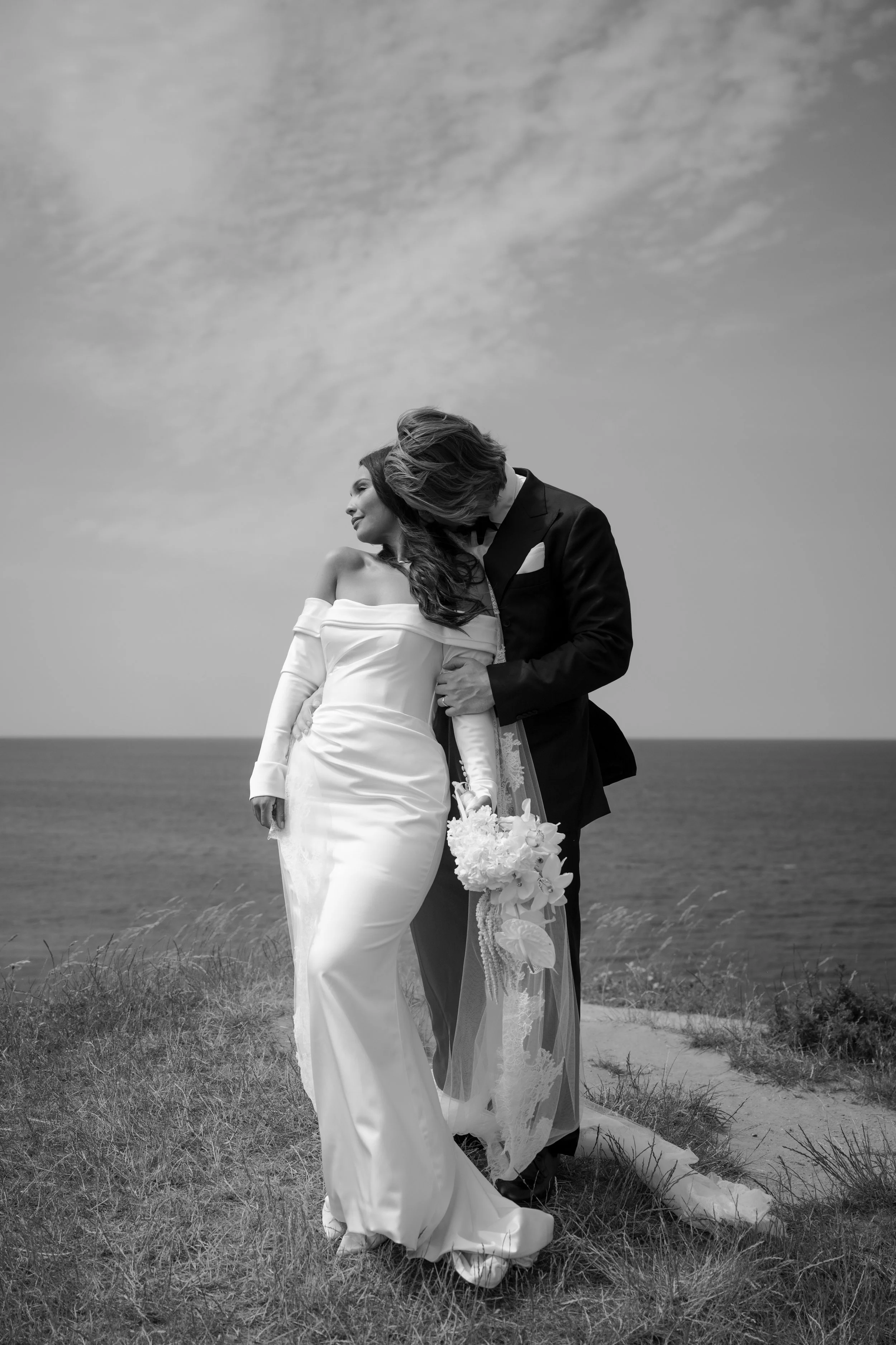 Black and white photo of a bride and groom kissing on a grassy hill with the ocean in the background.
