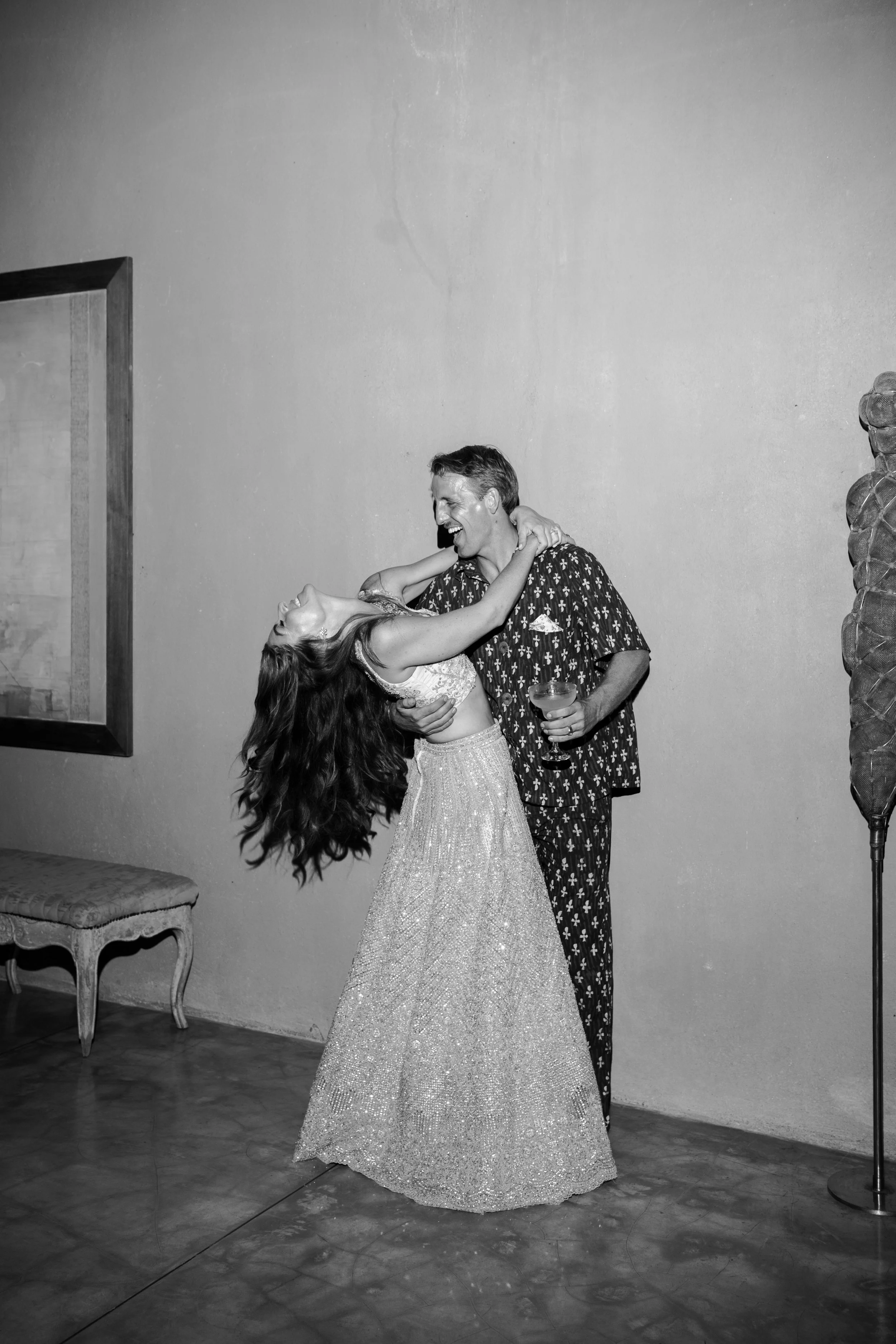 A woman in a sparkly dress and a man in patterned pajama-style shirt and pants dancing and laughing together indoors, holding drinks.