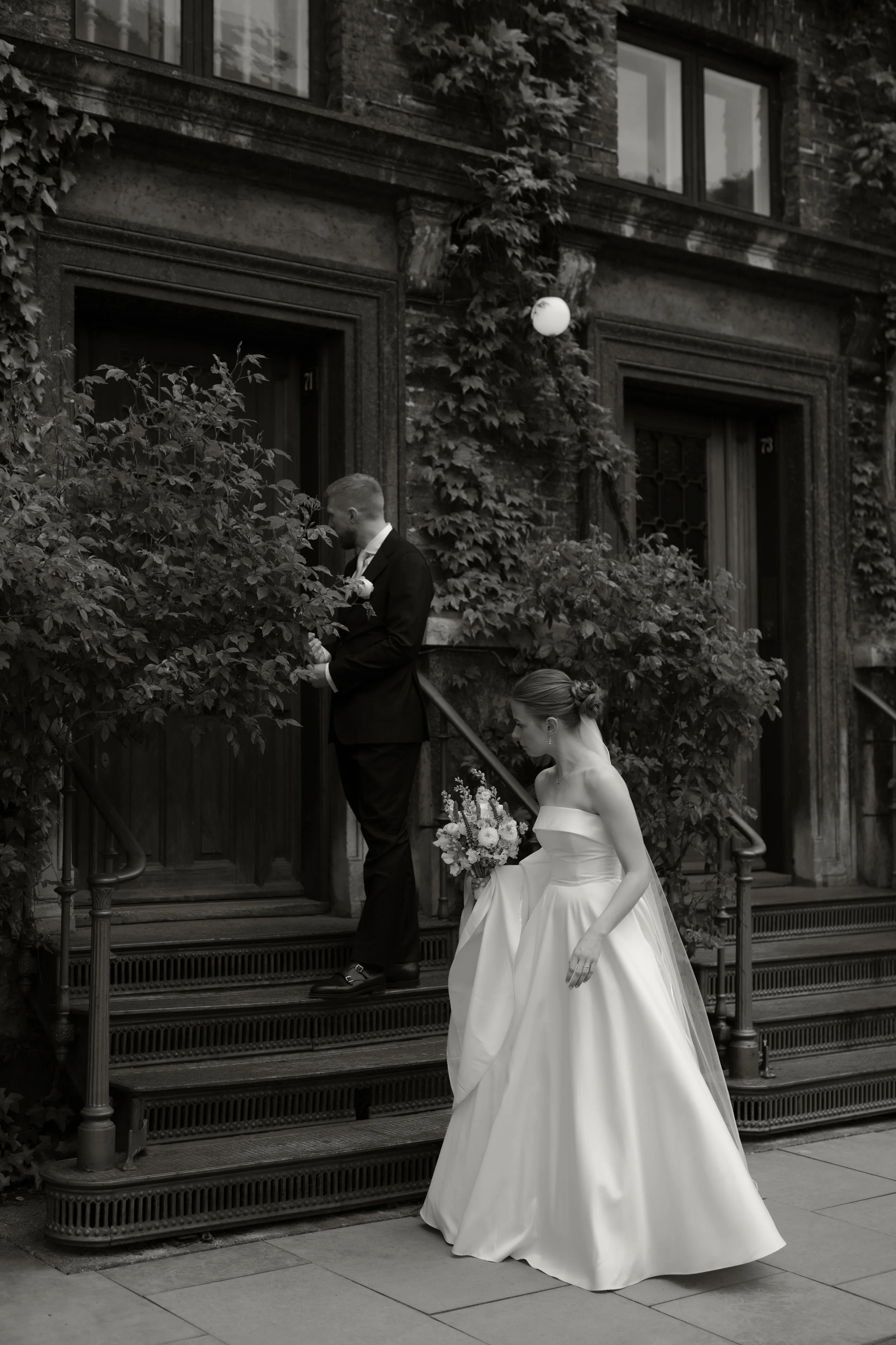 A black-and-white photo of a bride and groom outside a building, with the groom standing on stairs near the door and the bride holding a bouquet and walking down the stairs.