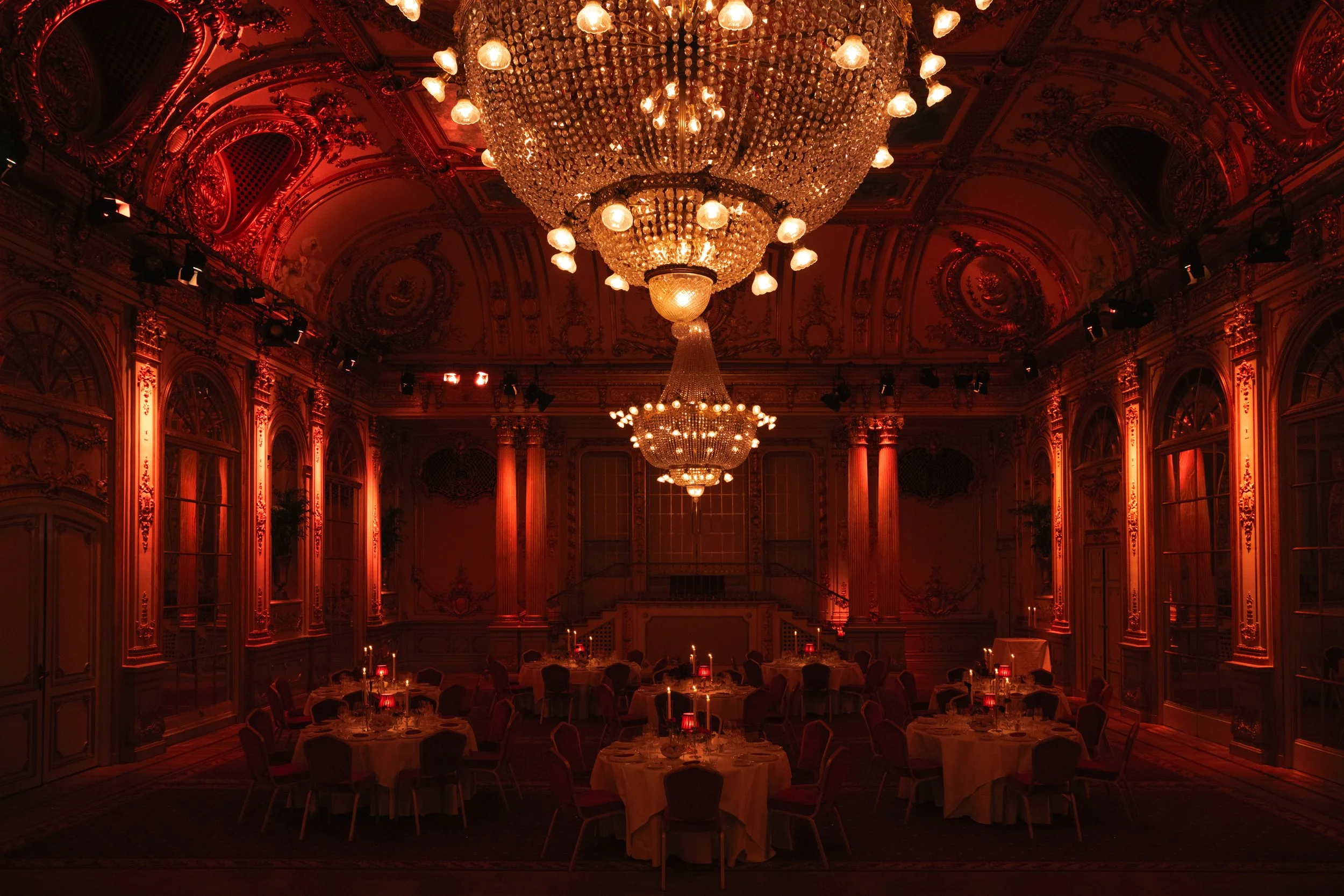 Elegant banquet hall with ornate gold and red decor, large crystal chandeliers, and round tables with white tablecloths and red cloth napkins, set for a formal event.