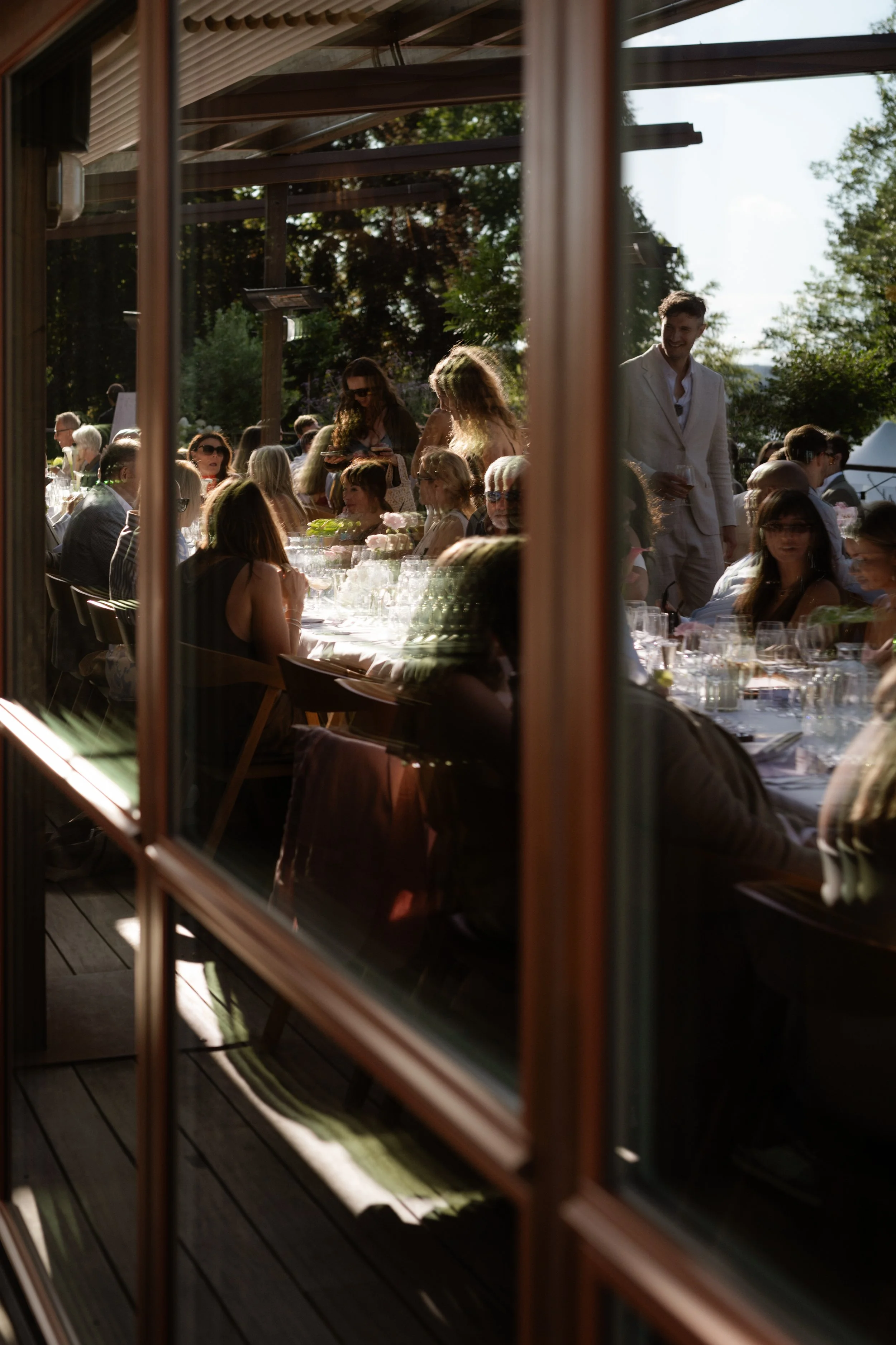 People gathered at an outdoor event, seen through a window with wooden frames, with food and drinks on the tables and trees in the background.