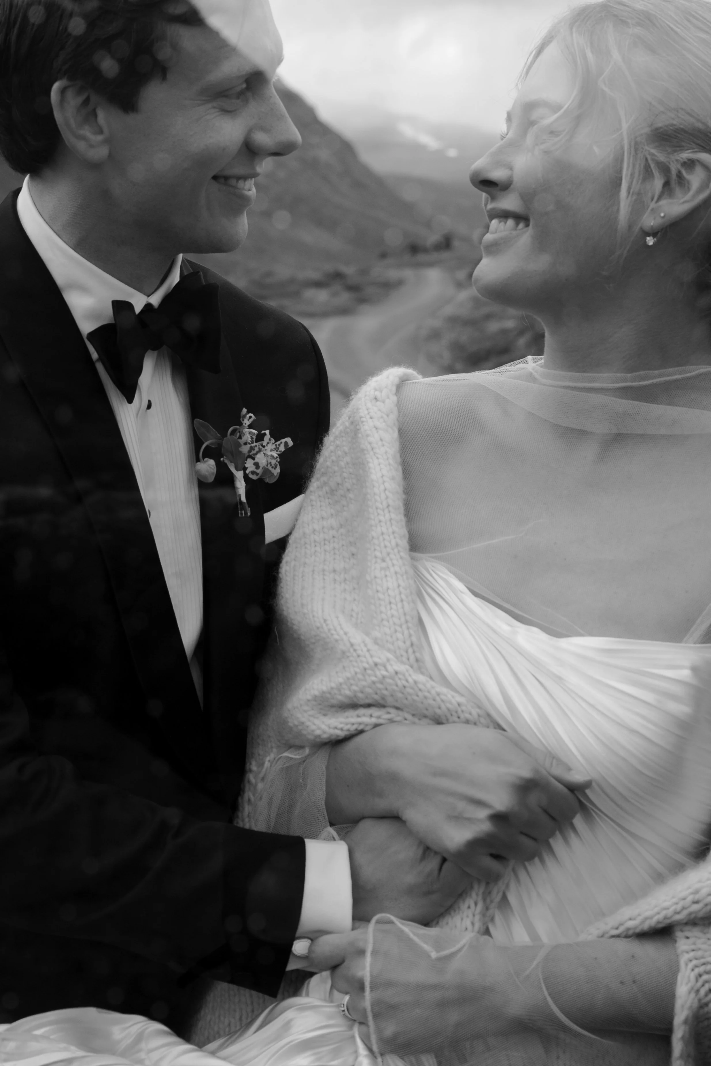 A black and white photo of a bride and groom smiling at each other outdoors, holding hands, with mountains and a winding road in the background.