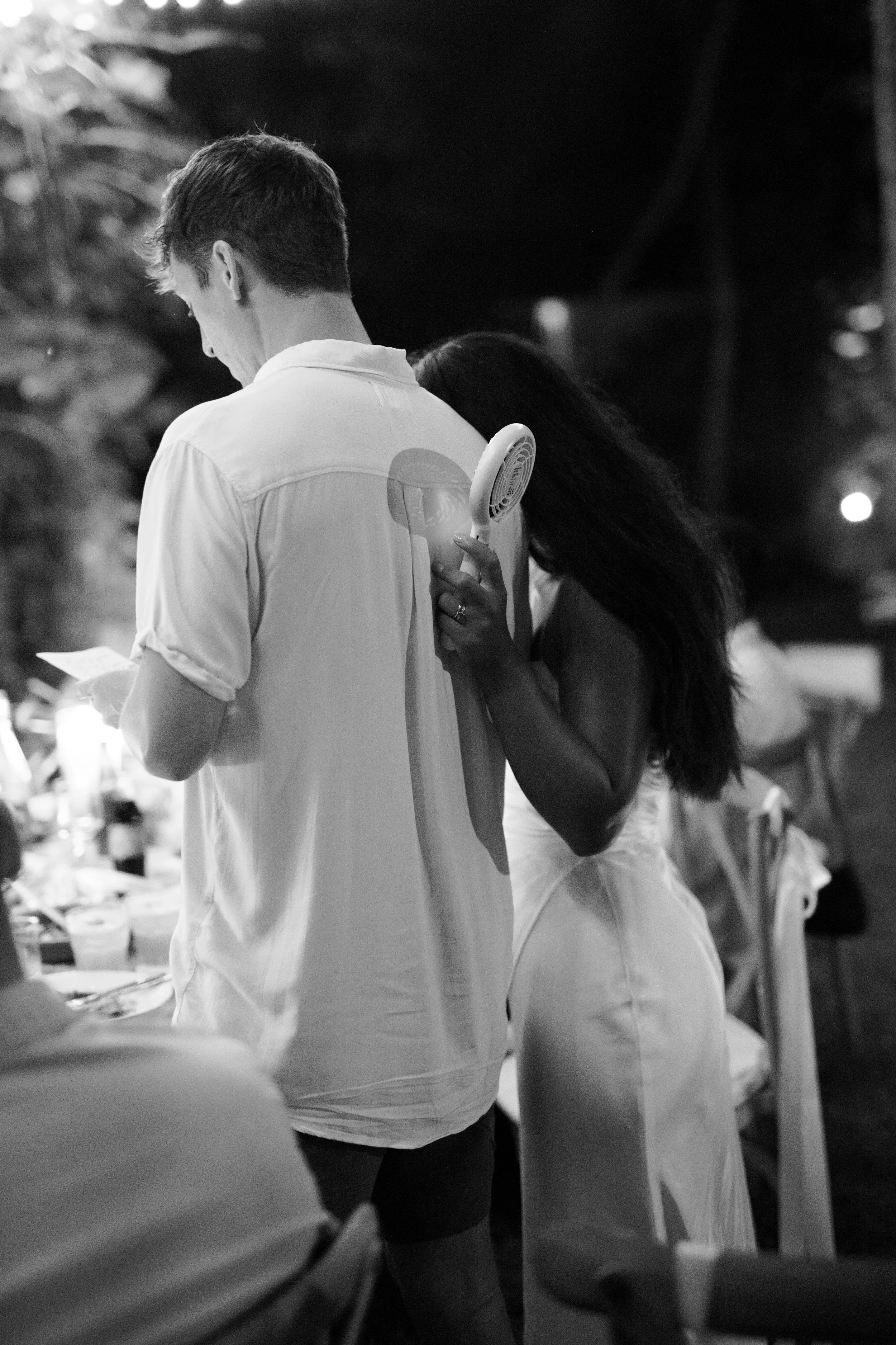 A woman is leaning on a man's back while holding a small handheld fan near his shoulder at an outdoor evening gathering.