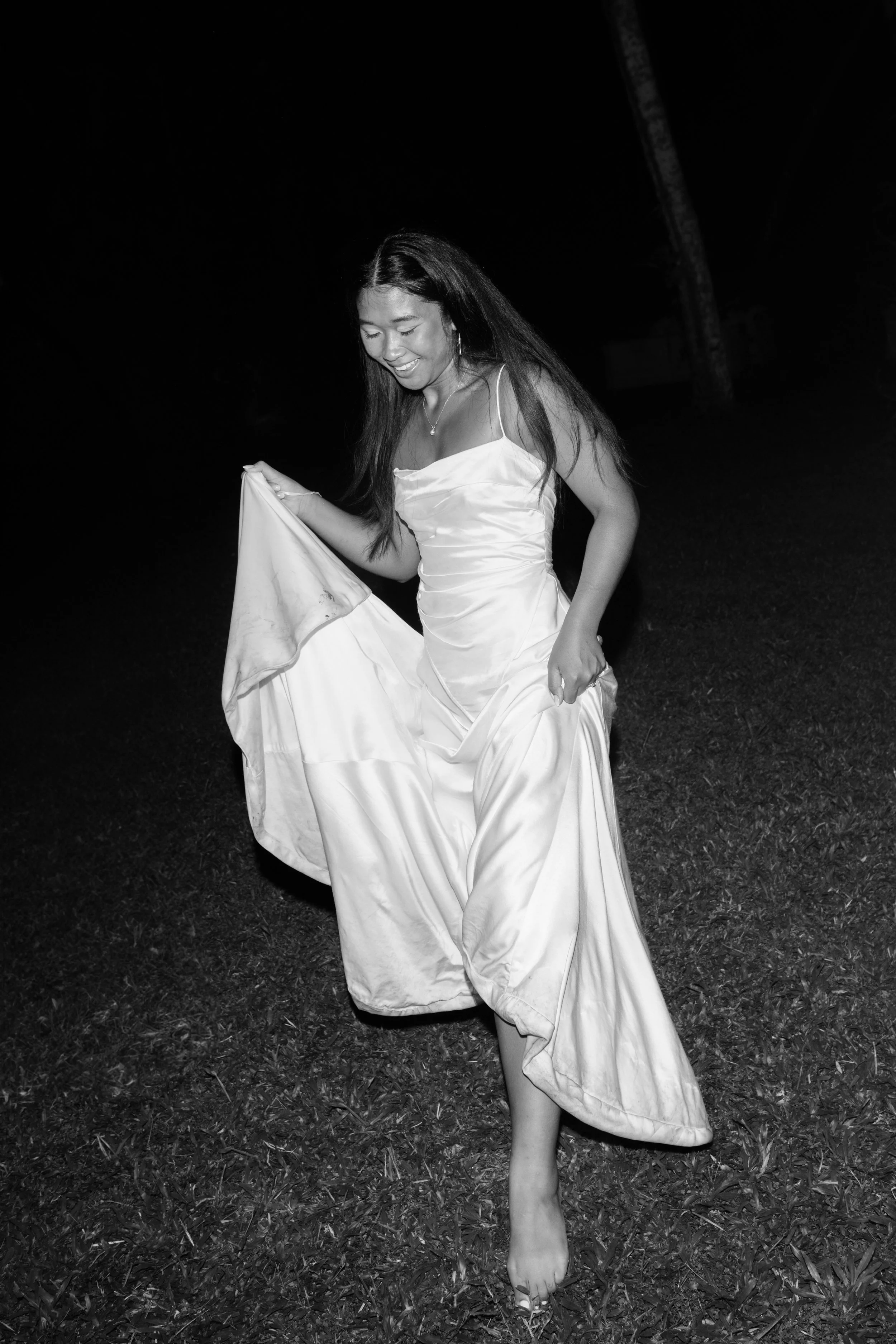 A young woman in a satin gown holding her dress while smiling, standing barefoot on grass at night.