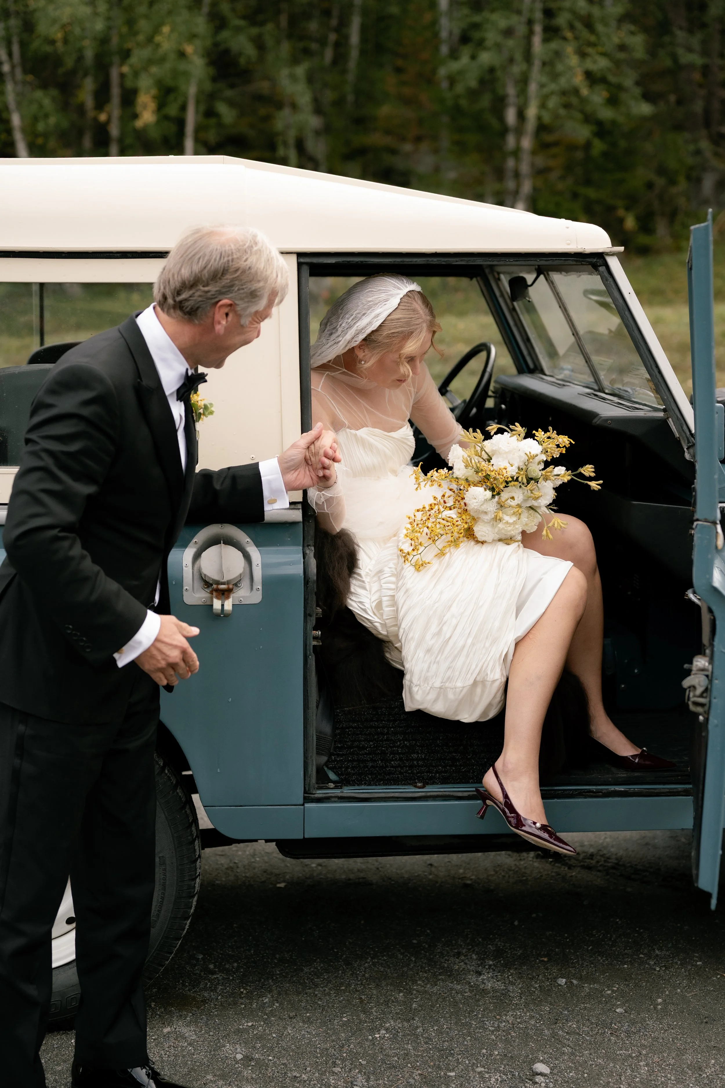 A bride sitting in a vintage car after her wedding, holding a bouquet of flowers, while a man in a tuxedo helps her get out of the vehicle.
