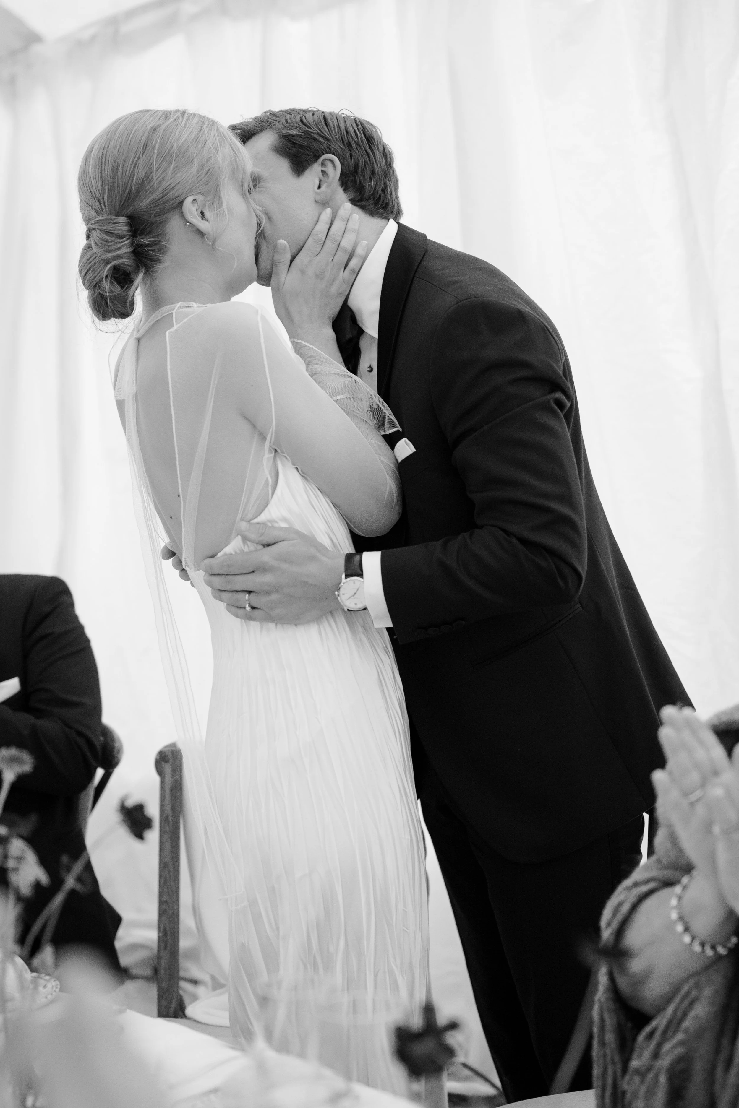 A couple is kissing at their wedding ceremony, with the woman in a wedding dress and the man in a suit, indoors with white curtains.