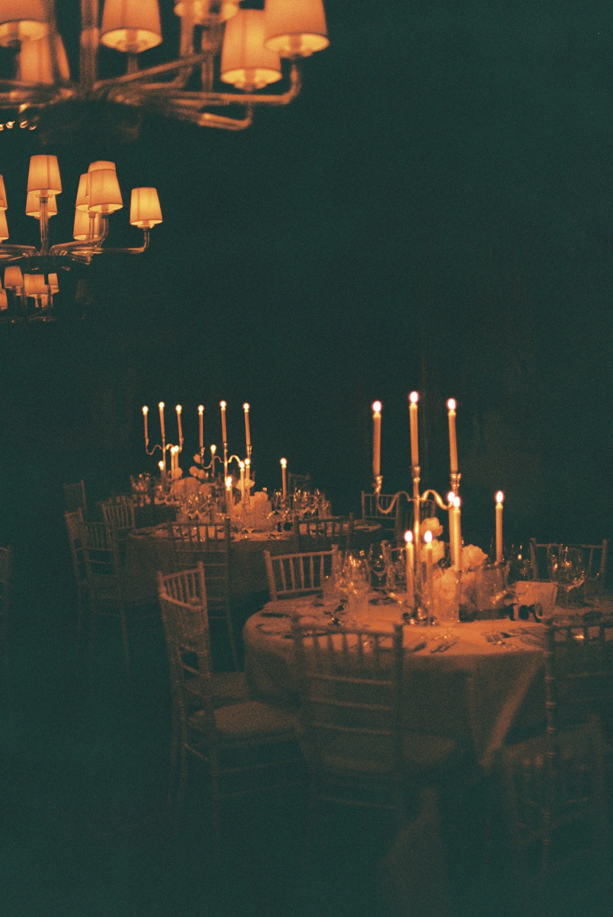 A dimly lit dining room with round tables, tall candles, and elegant glassware, set for a formal event.