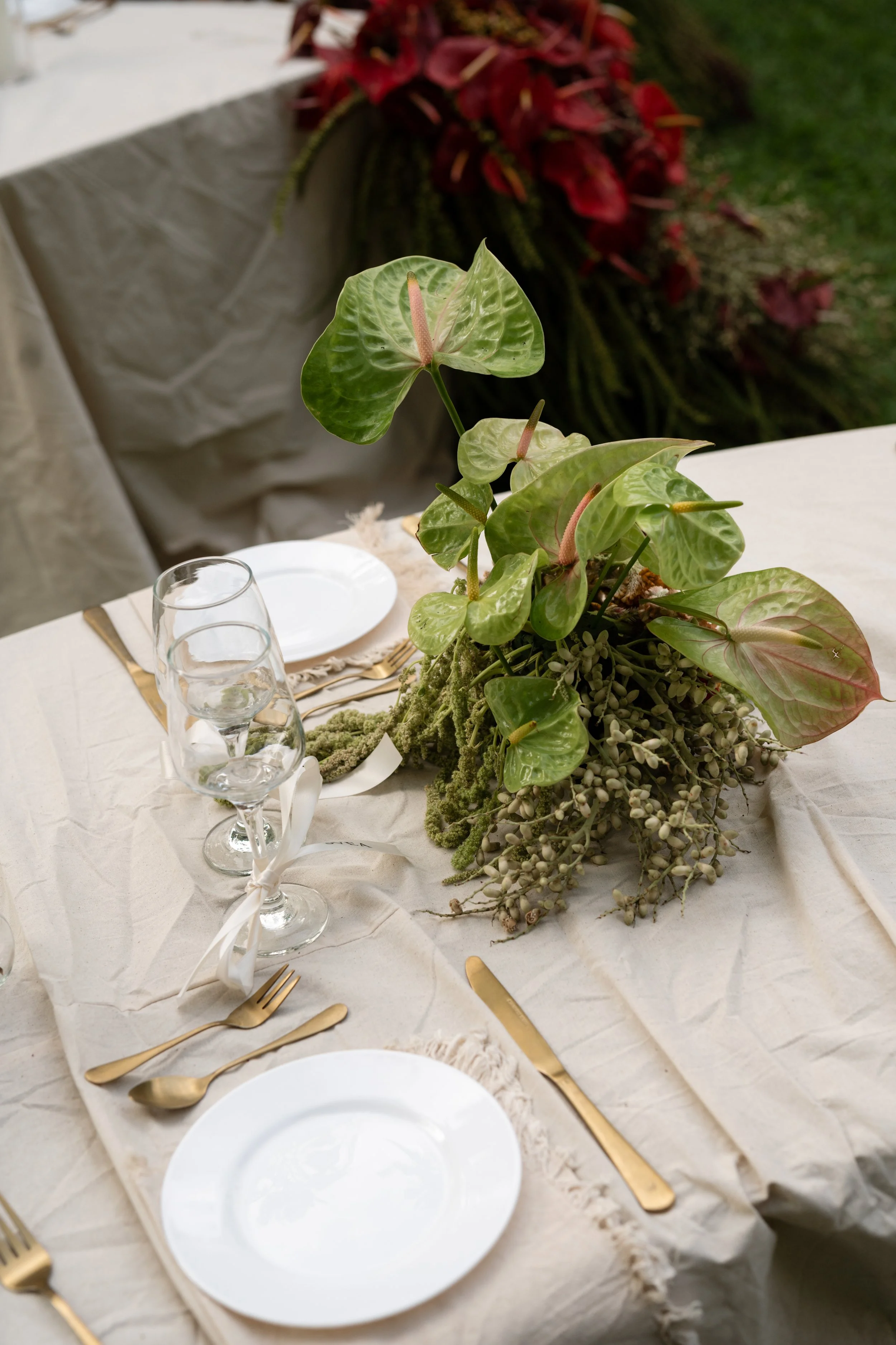 Table setting with white plates, gold utensils, water glasses, and a centerpiece of green leafy plants and hanging greenery.
