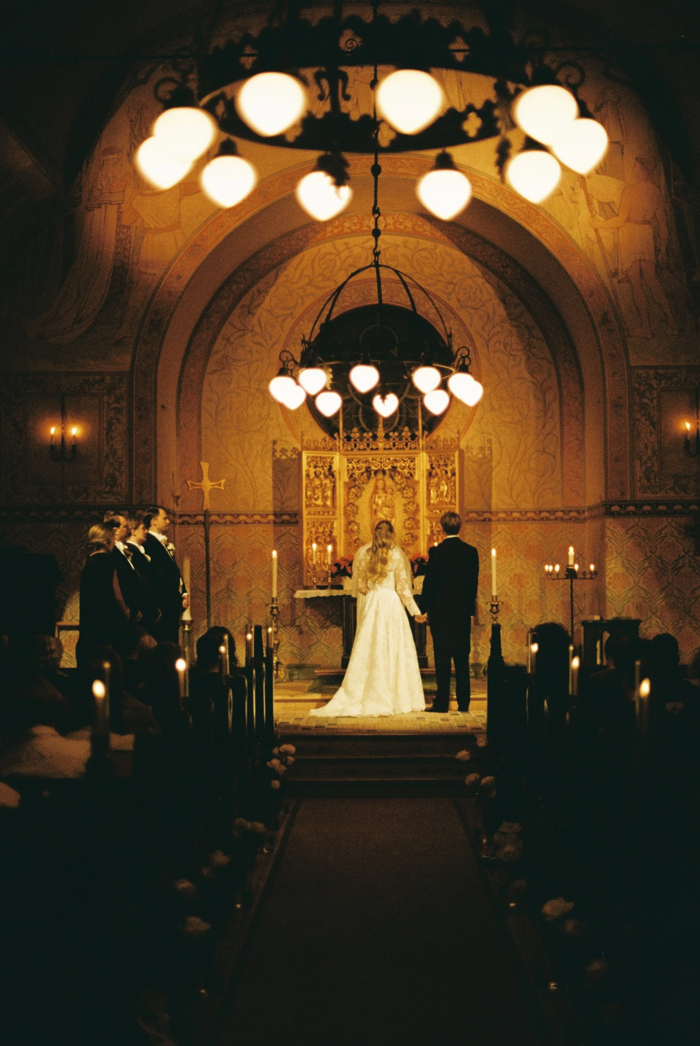 A wedding ceremony taking place inside a church, with a bride and groom holding hands at the altar, surrounded by bridesmaids and groomsmen, under chandeliers and candlelight.