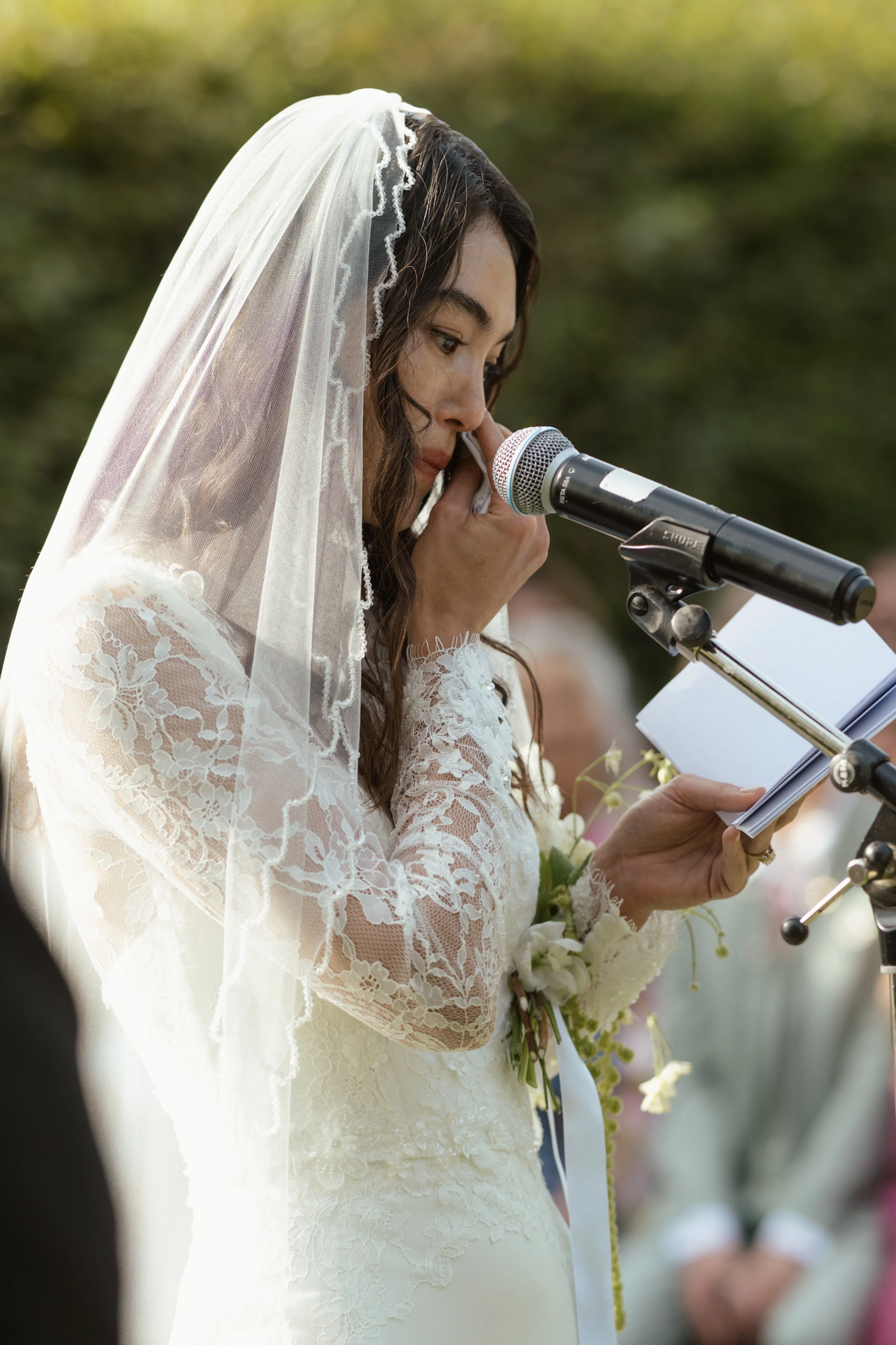 A bride in a lace wedding dress and veil reading vows into a microphone during an outdoor wedding ceremony.