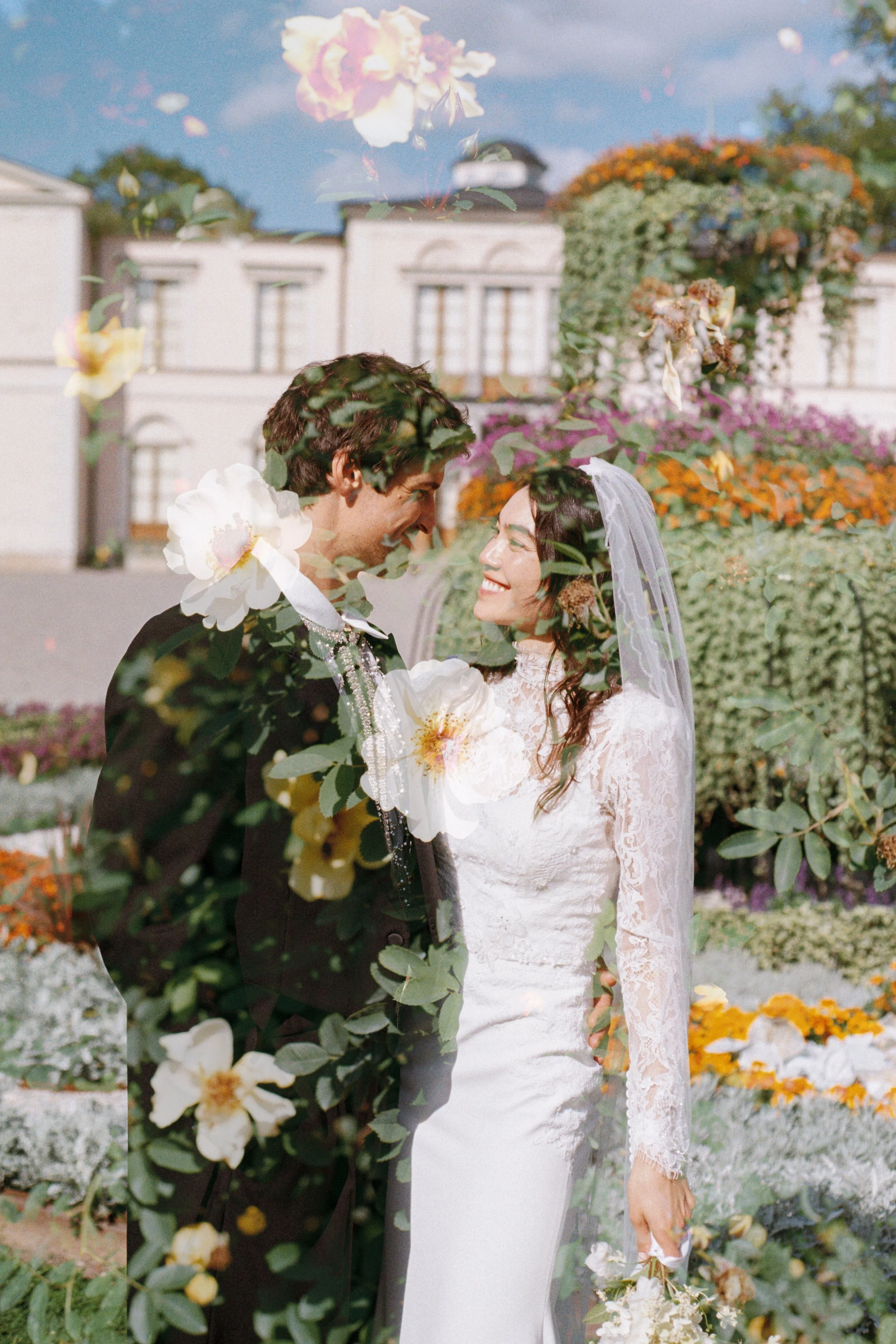 A bride and groom smiling at each other in a garden with colorful flowers, with a building in the background.