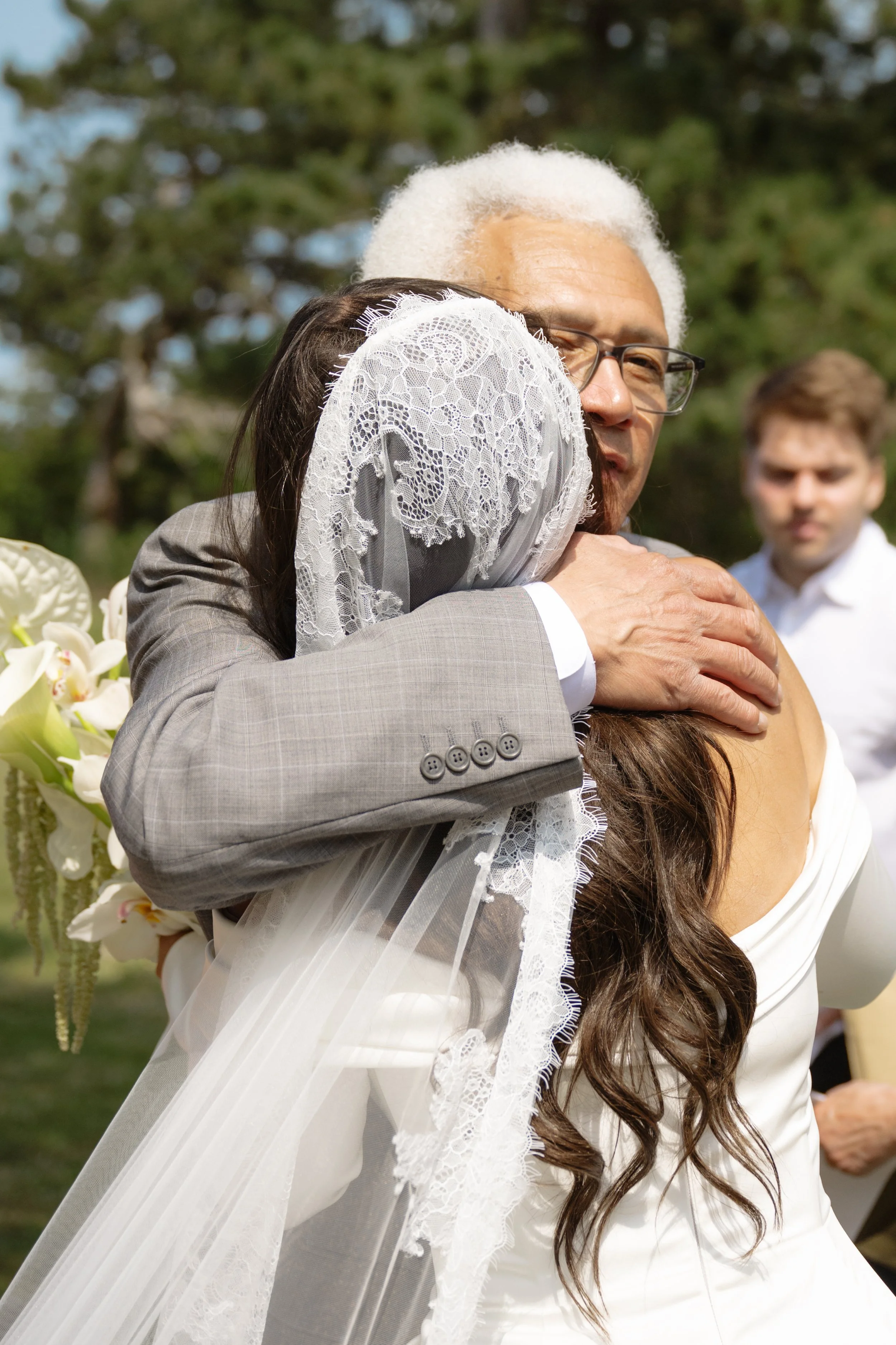 A wedding hug between an elderly man with white hair and glasses and a bride wearing a lace veil and white dress, with a young man in a white shirt in the background outside.