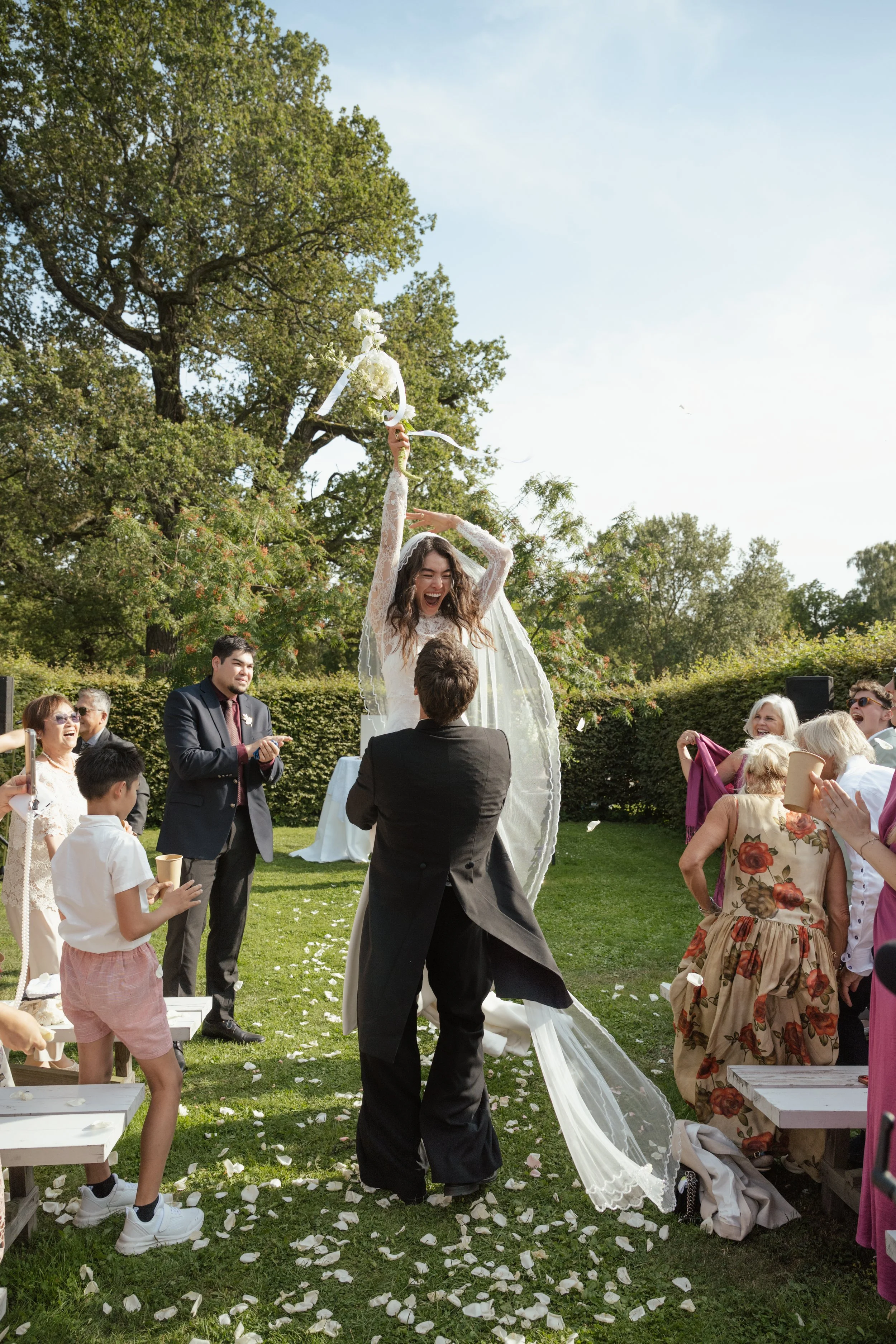 Bride holding a bouquet of white flowers while jumping in the air during an outdoor wedding celebration, surrounded by friends and family.