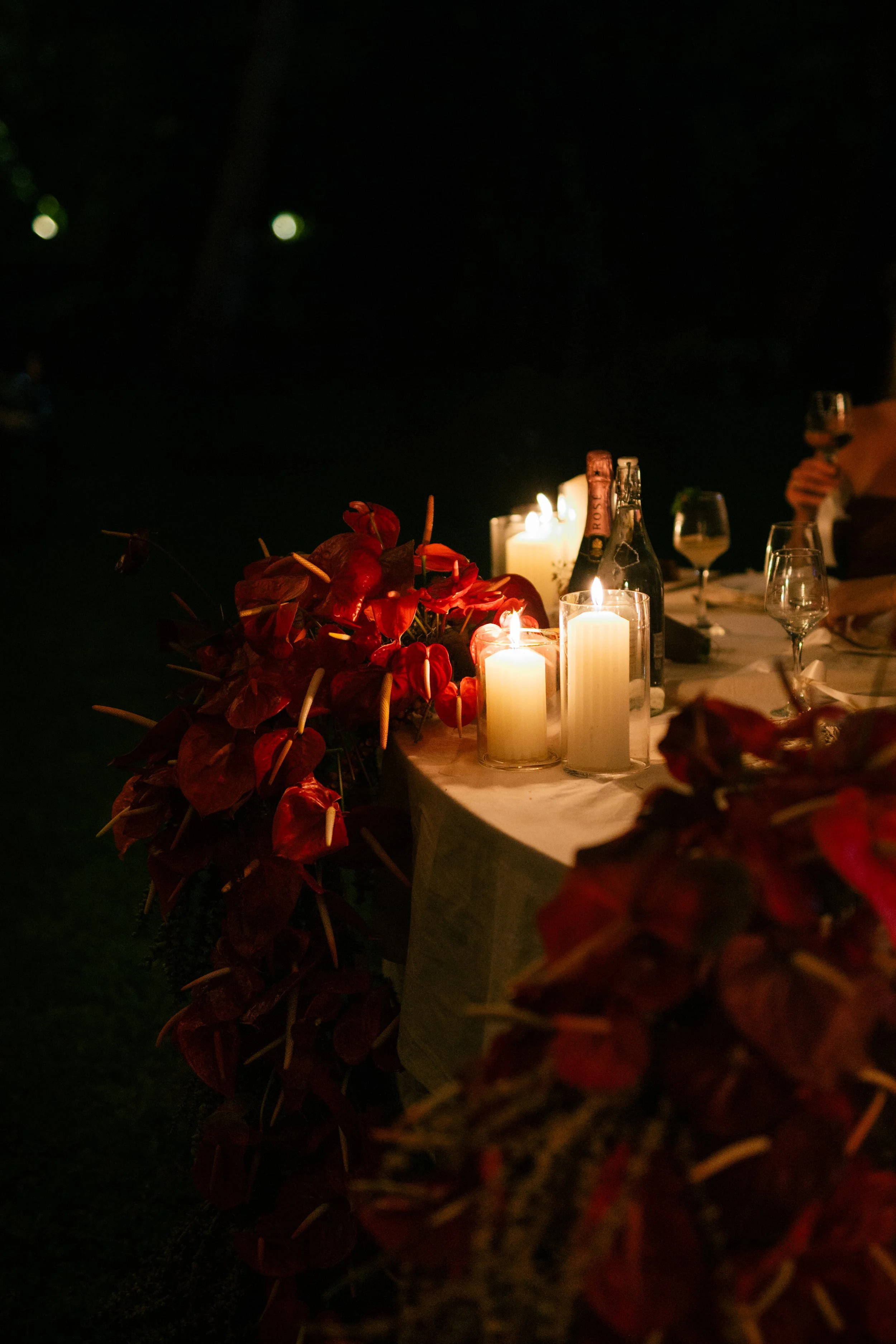 A decorated table with white candles and red flowers at night, with wine glasses and bottles.