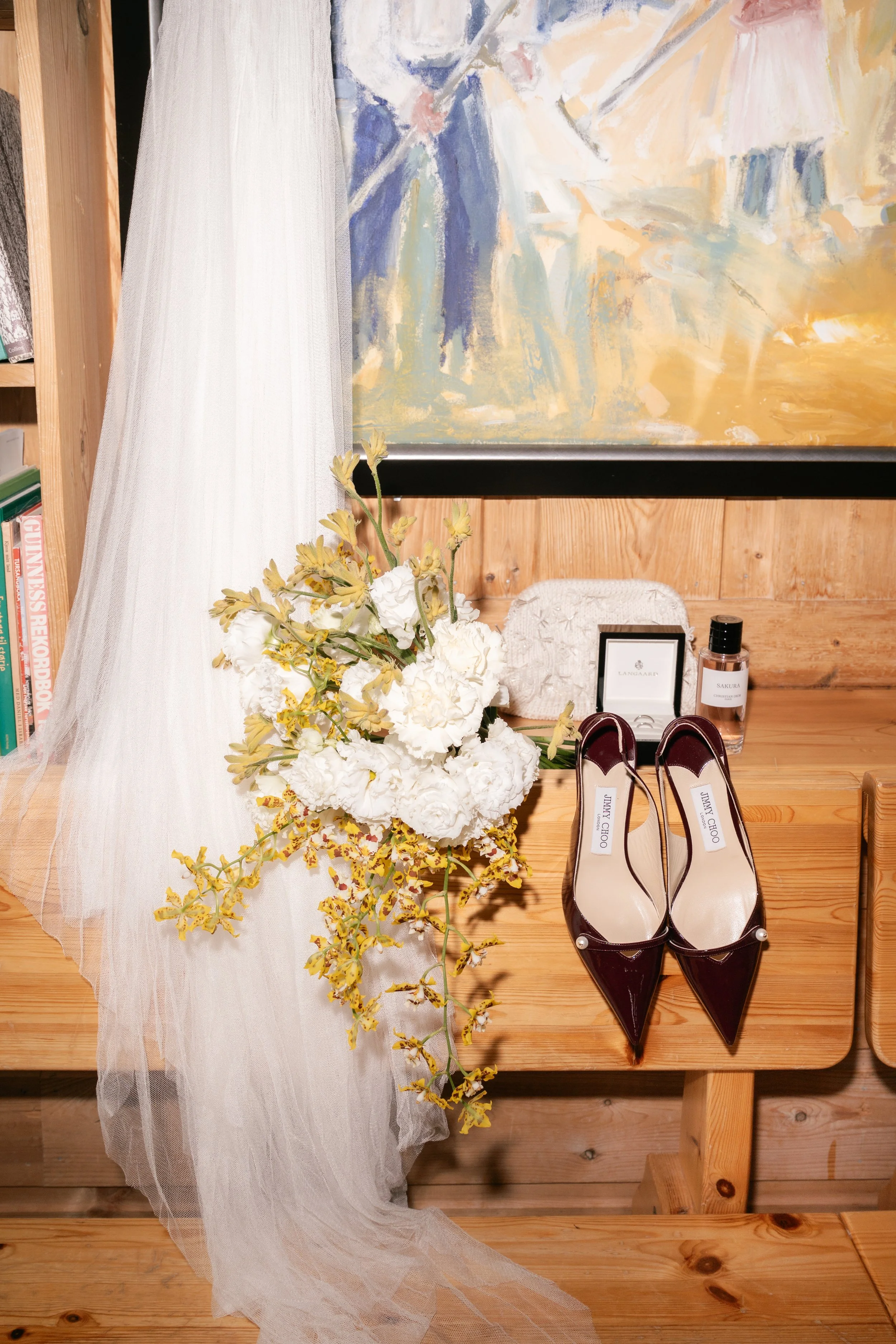 A wooden table with a bouquet of white flowers, a white purse, a pair of burgundy Jimmy Choo high heels, a framed photo, a perfume bottle, and a bottle of sakura fragrance. A sheer white curtain and an abstract painting are in the background.