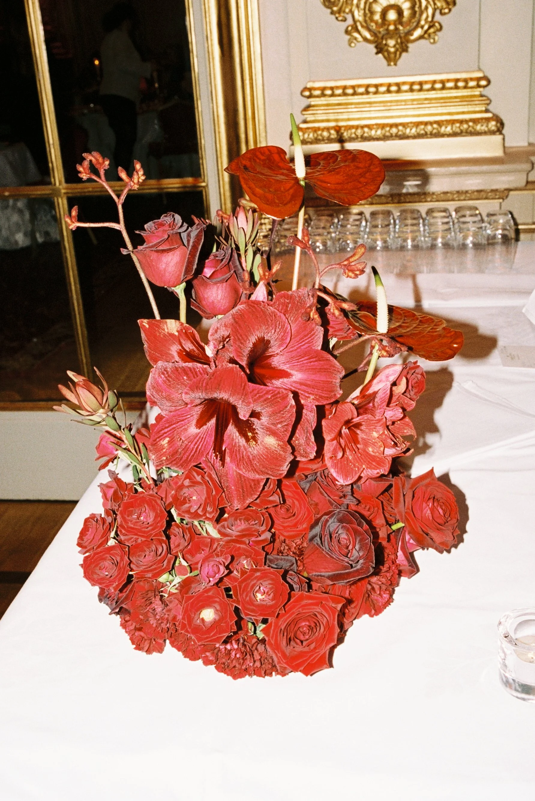 A floral arrangement featuring red roses, lilies, anthuriums, and other flowers on a white table, with a mirror and decorative gold accents in the background.