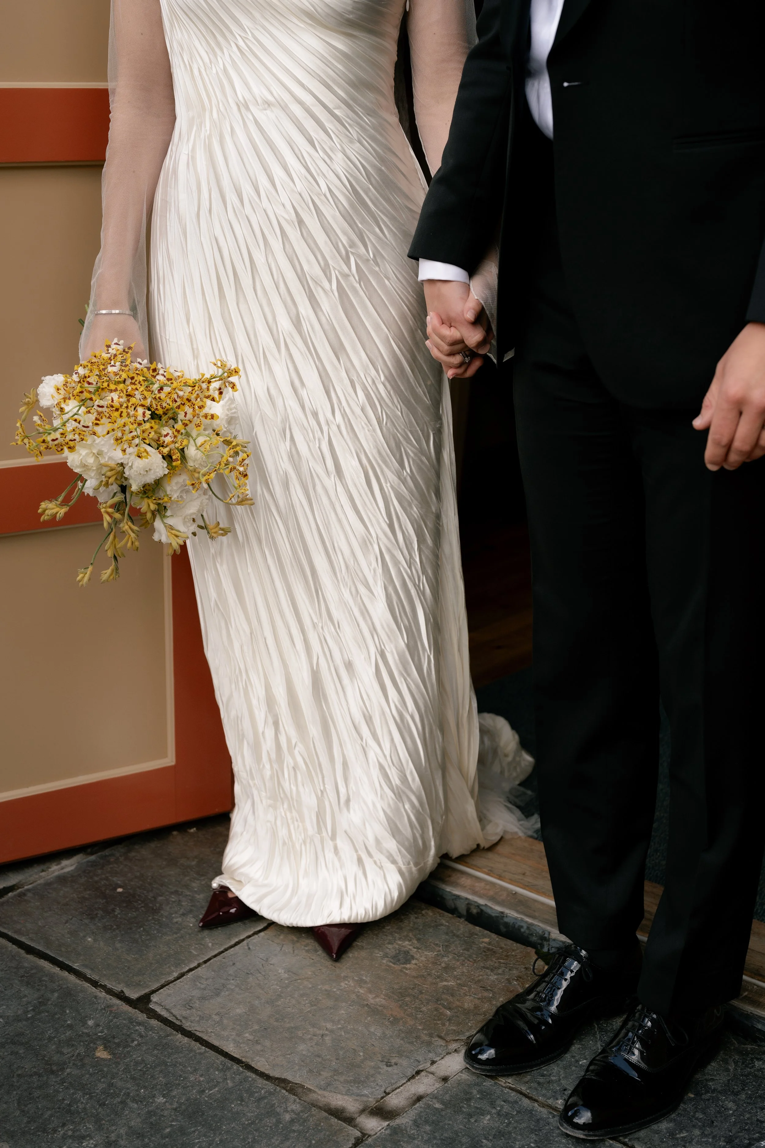 Bride and groom holding hands during their wedding ceremony, with the bride wearing a white dress and holding a bouquet, and the groom in black tuxedo.