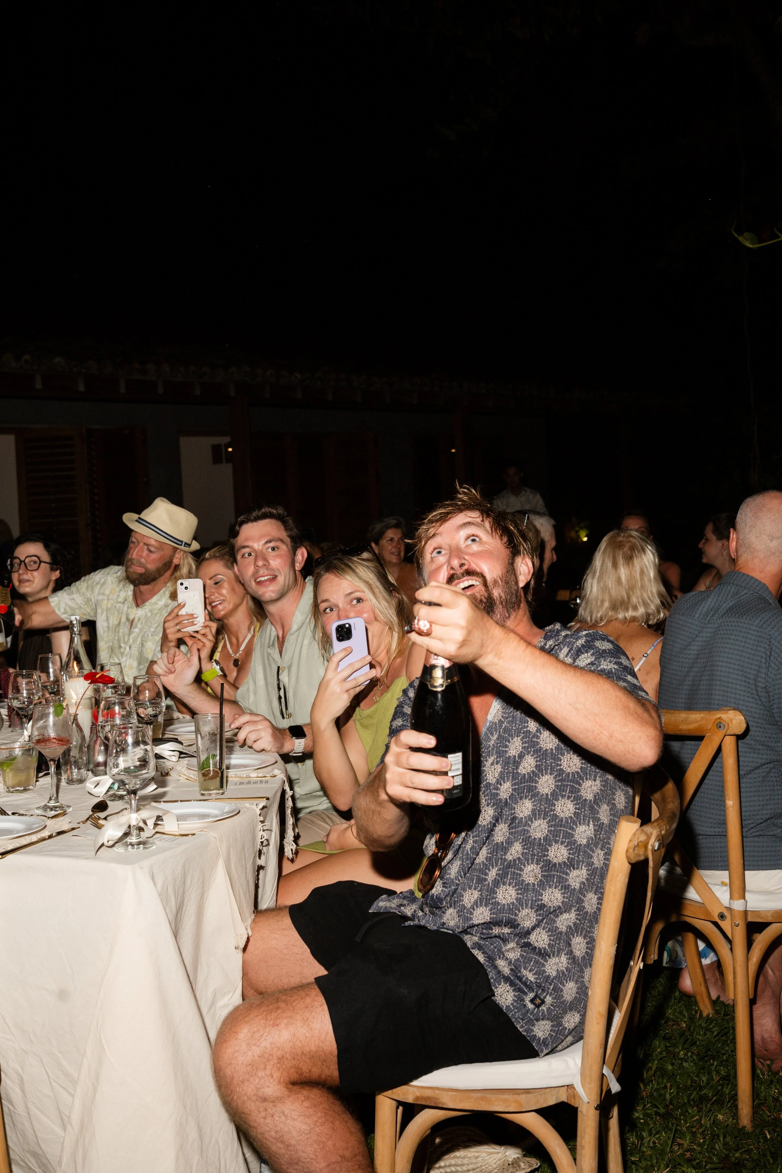 People sitting at a dinner table, some are taking photos or videos, and one man is opening a bottle of champagne at a celebration or party.