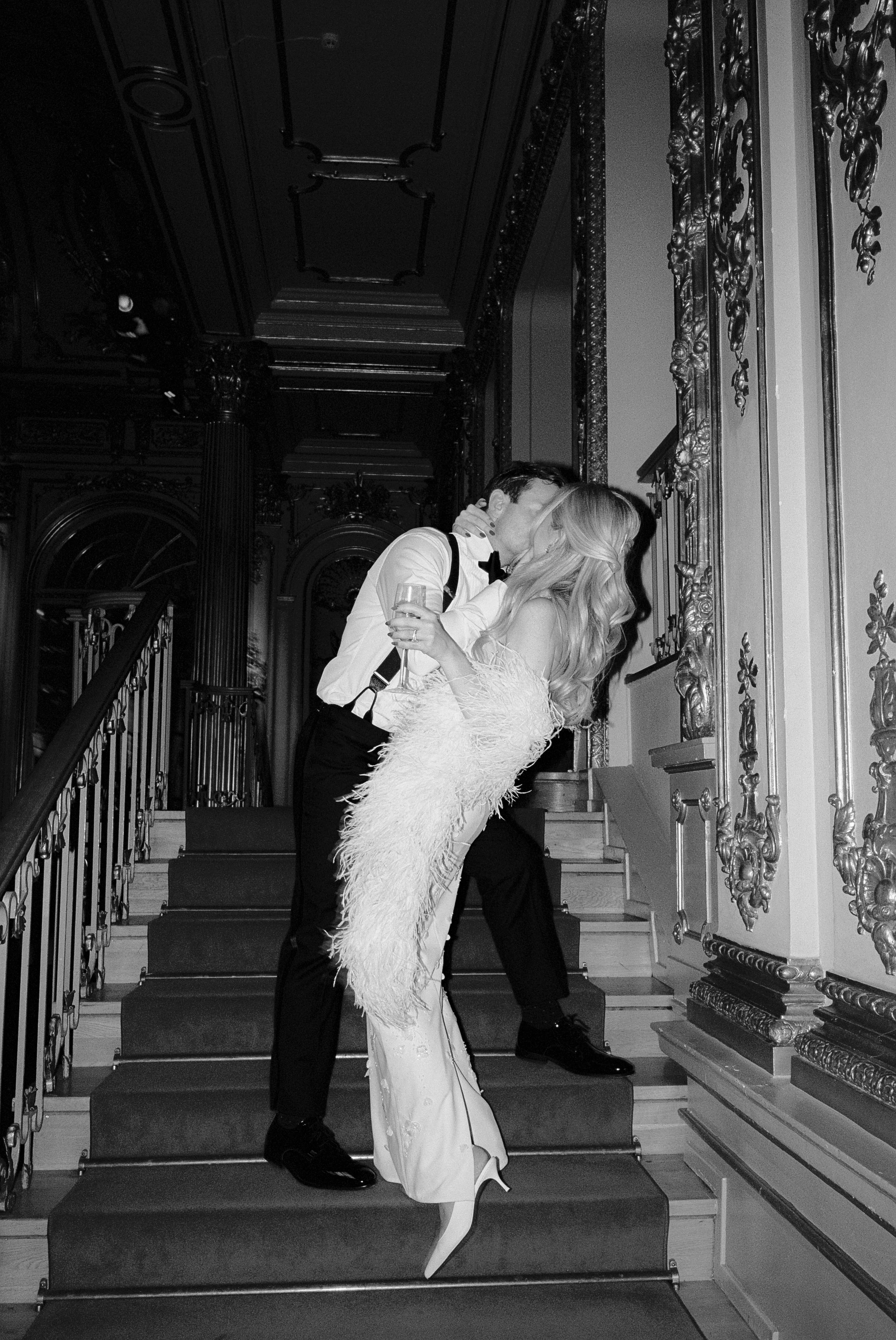 A black and white photo of a couple kissing on a grand staircase, dressed in formal attire, with the woman holding a glass of champagne.