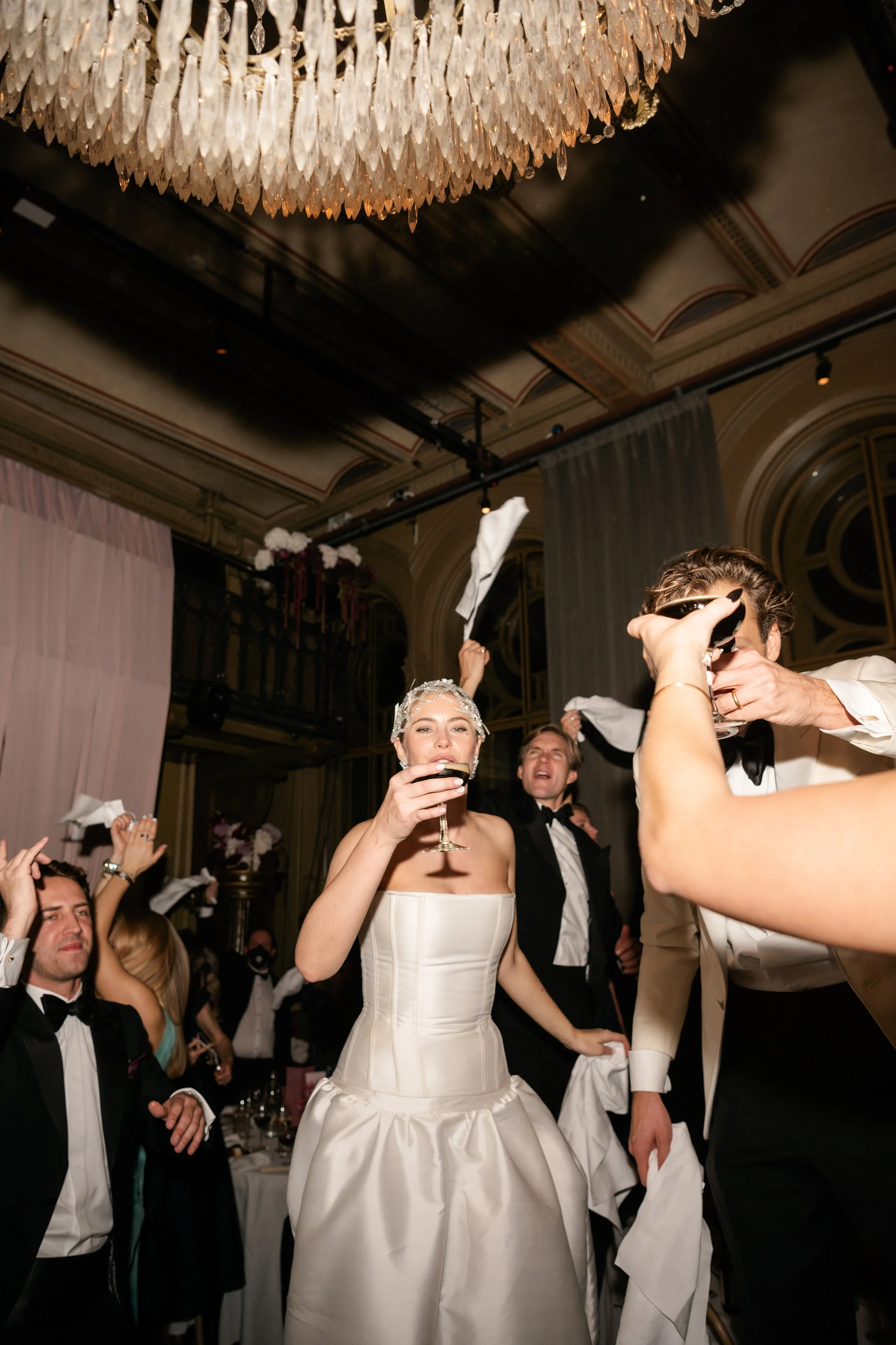 A bride in a white wedding dress holds a glass while celebrating with guests at a wedding reception inside a decorated venue with a chandelier and ornate ceiling.