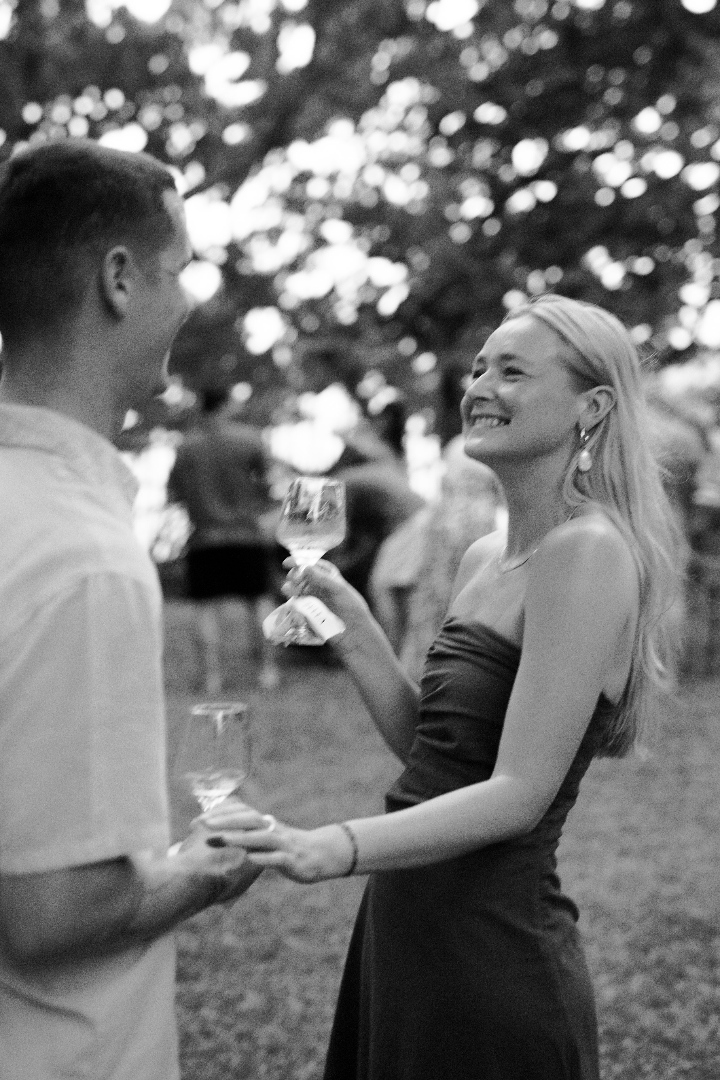 A woman in a strapless dress smiling and holding a glass of wine, engaging with a man outdoors at dusk, with trees in the background.