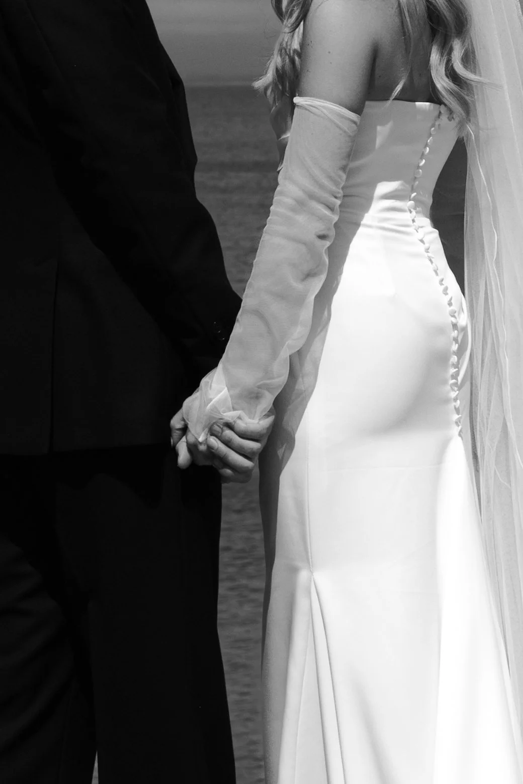 A black-and-white photo of a bride and groom holding hands during their wedding, with the bride in a white wedding dress and the groom in a dark suit.