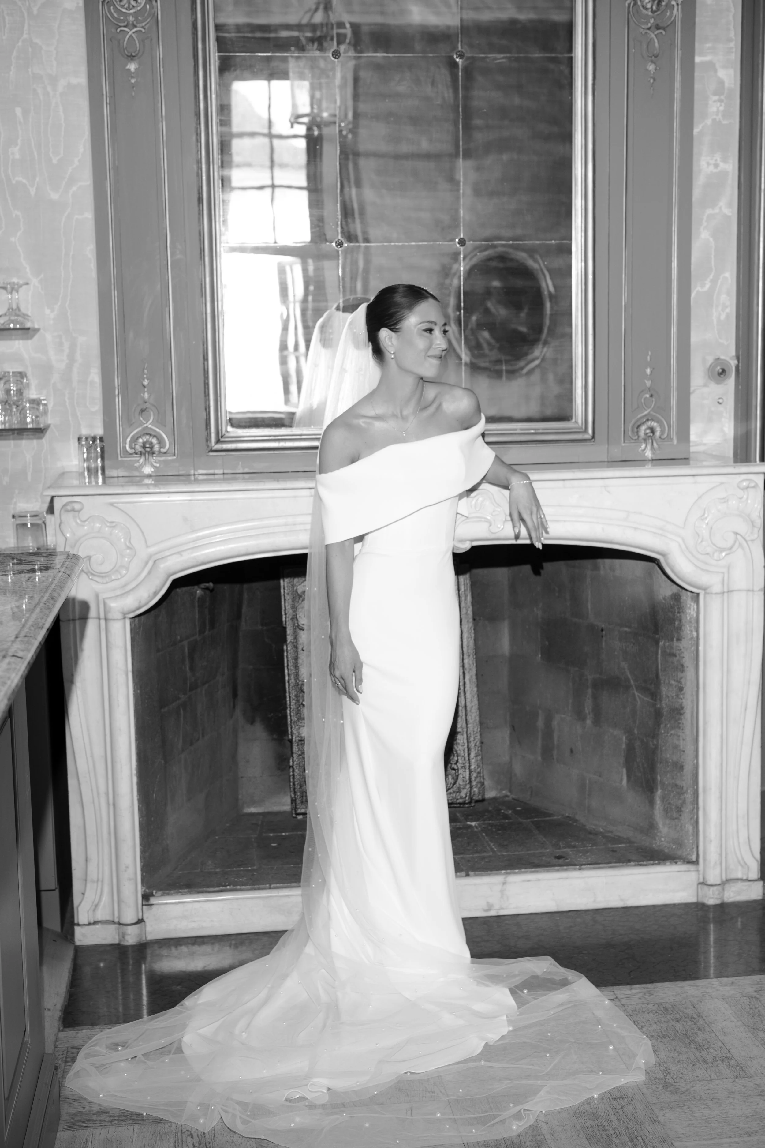 Bride in a strapless wedding dress with off-shoulder design and veil, standing in front of a fireplace and mirror, smiling softly.