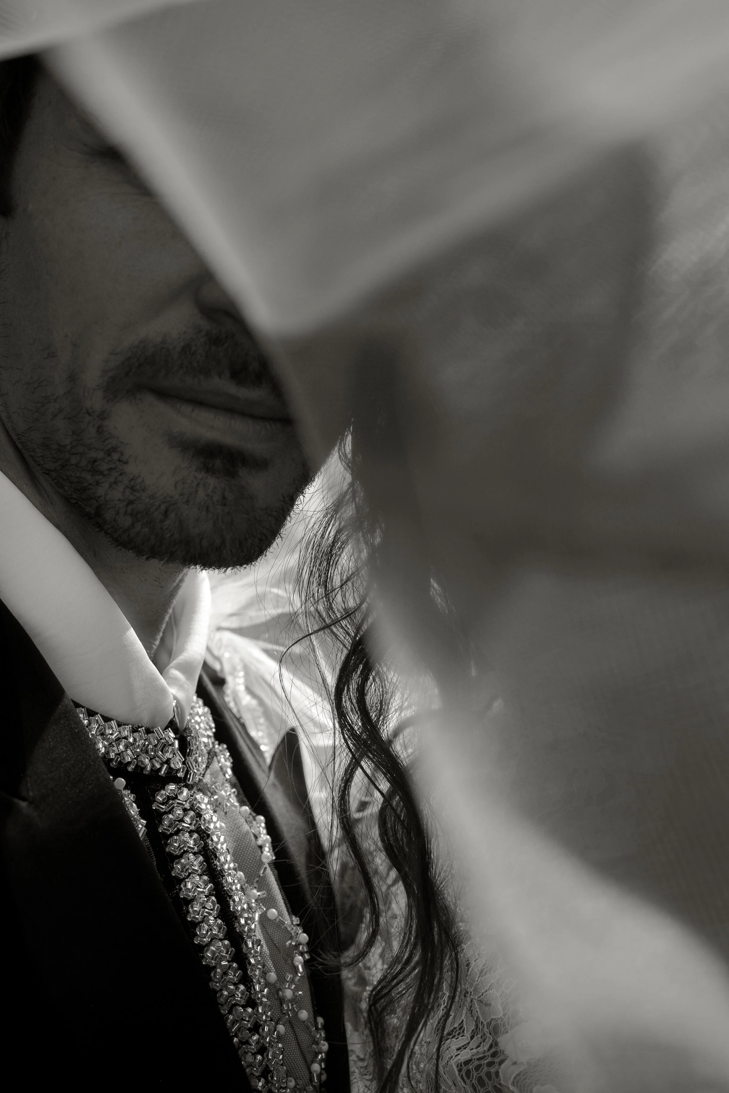 A black and white close-up photo of a man with a beard and mustache, partially obscured by a dramatic, flowing piece of fabric or curtain, with visible jewelry and lace details on his clothing.