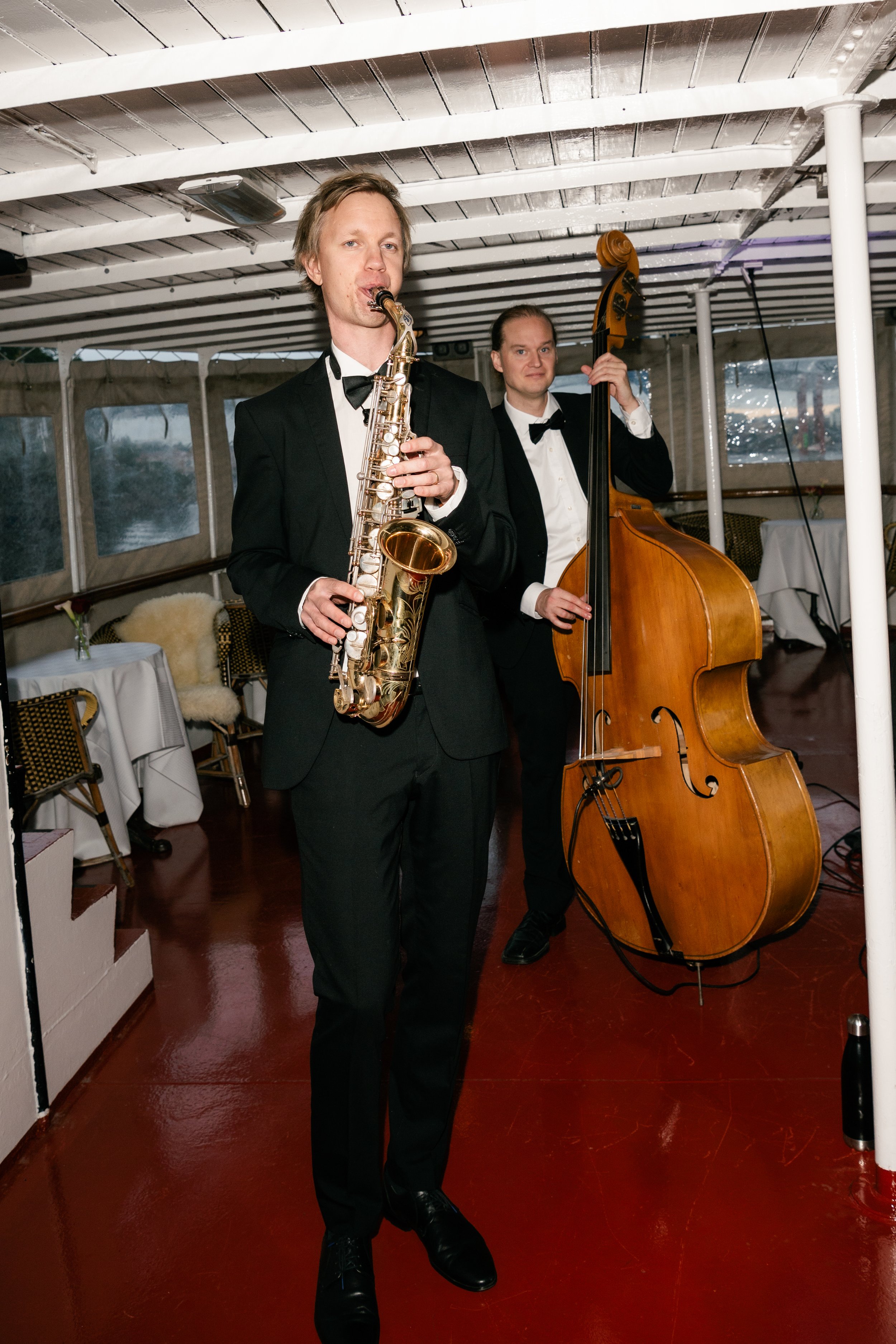 Two musicians in tuxedos perform, one playing a saxophone and the other playing a double bass, inside a decorated venue with tables and chairs.