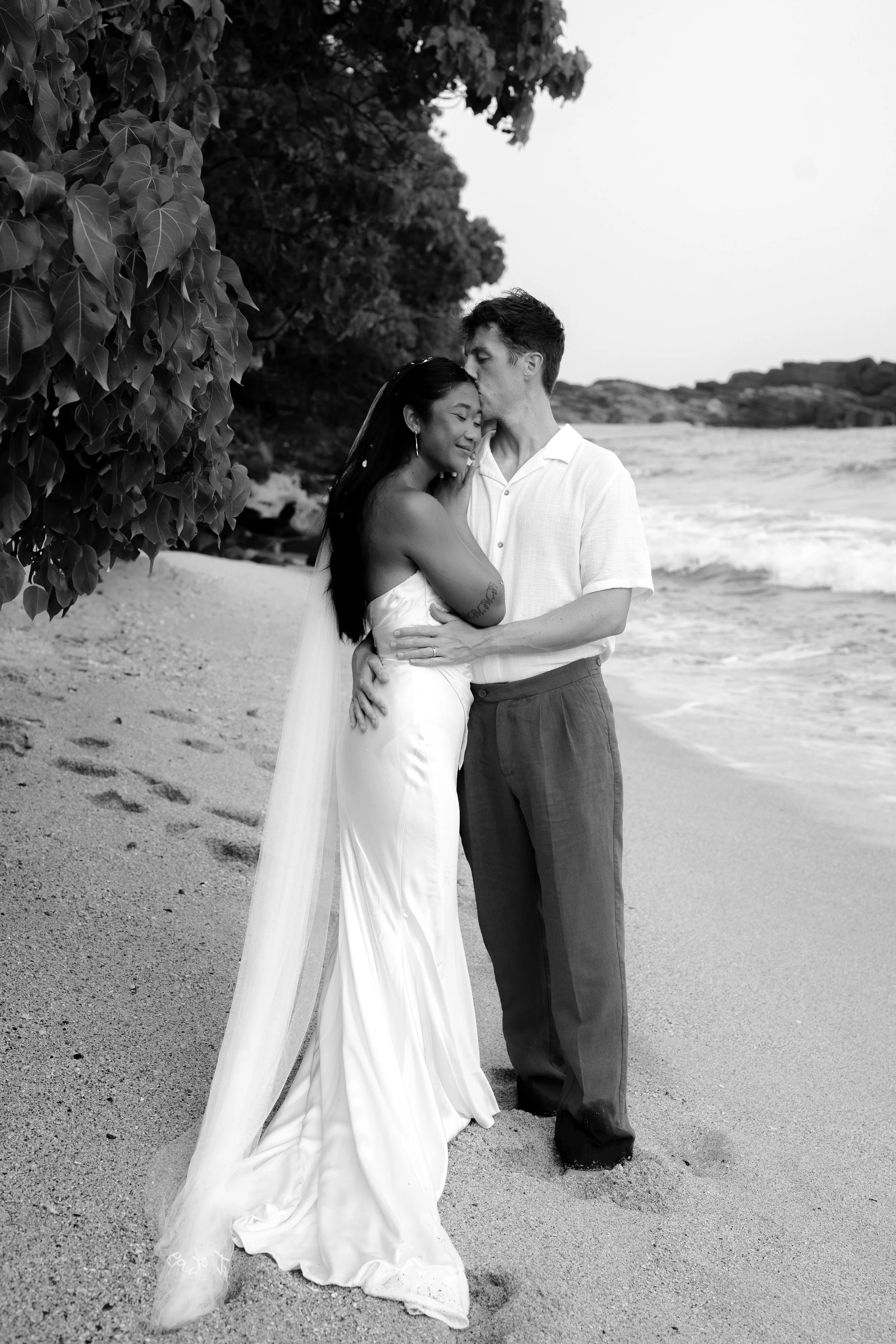 A couple stands on a sandy beach, embracing affectionately. The woman wears a strapless wedding gown with a long train, and the man wears a short-sleeved button-up shirt and trousers. They are near water with waves, trees, and rocks in the background, sharing an intimate moment.