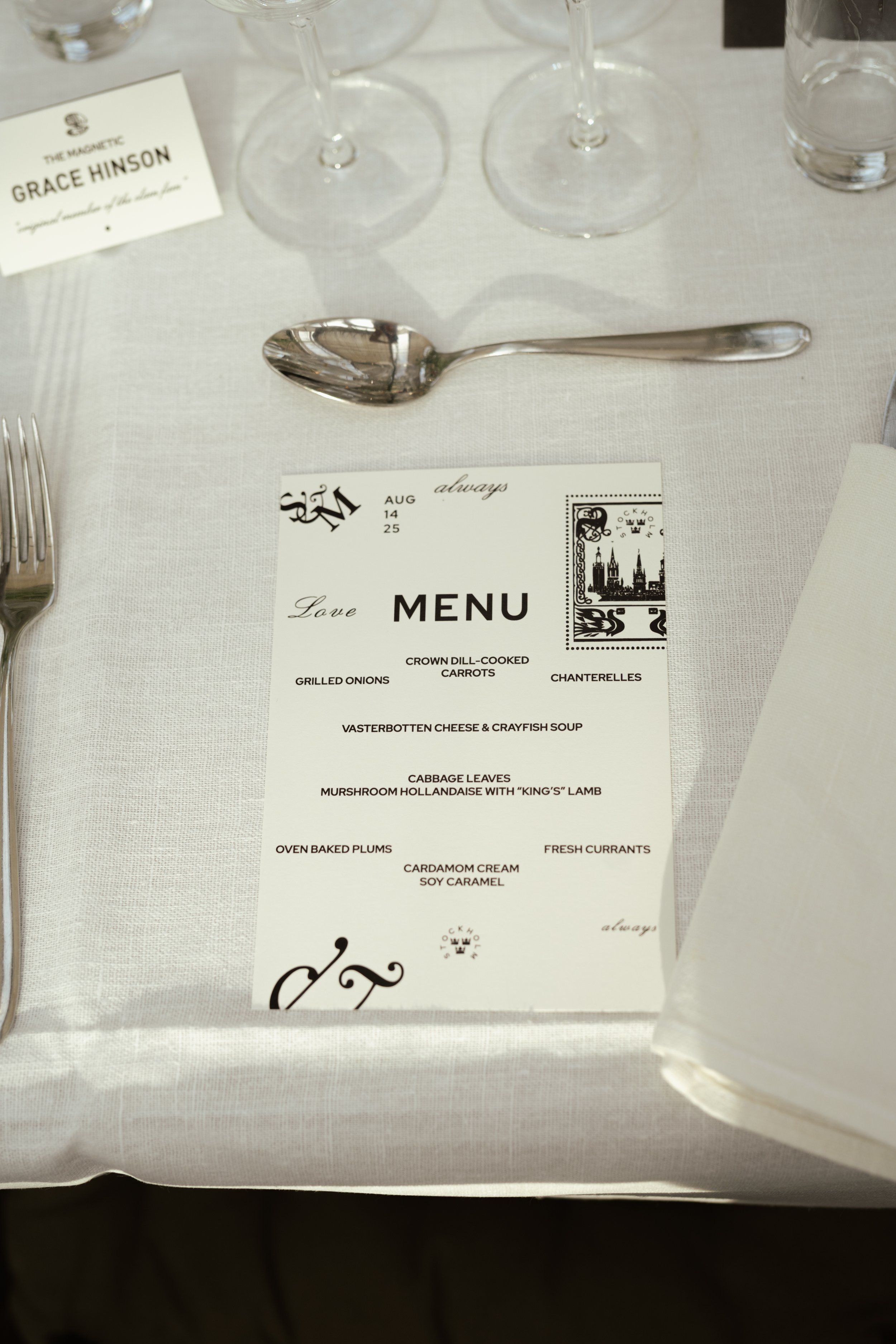 A wedding reception table setting with a menu card, a fork on the left, a spoon above the menu, wine glasses, a glass of water, and a place card for Grace Hinson.