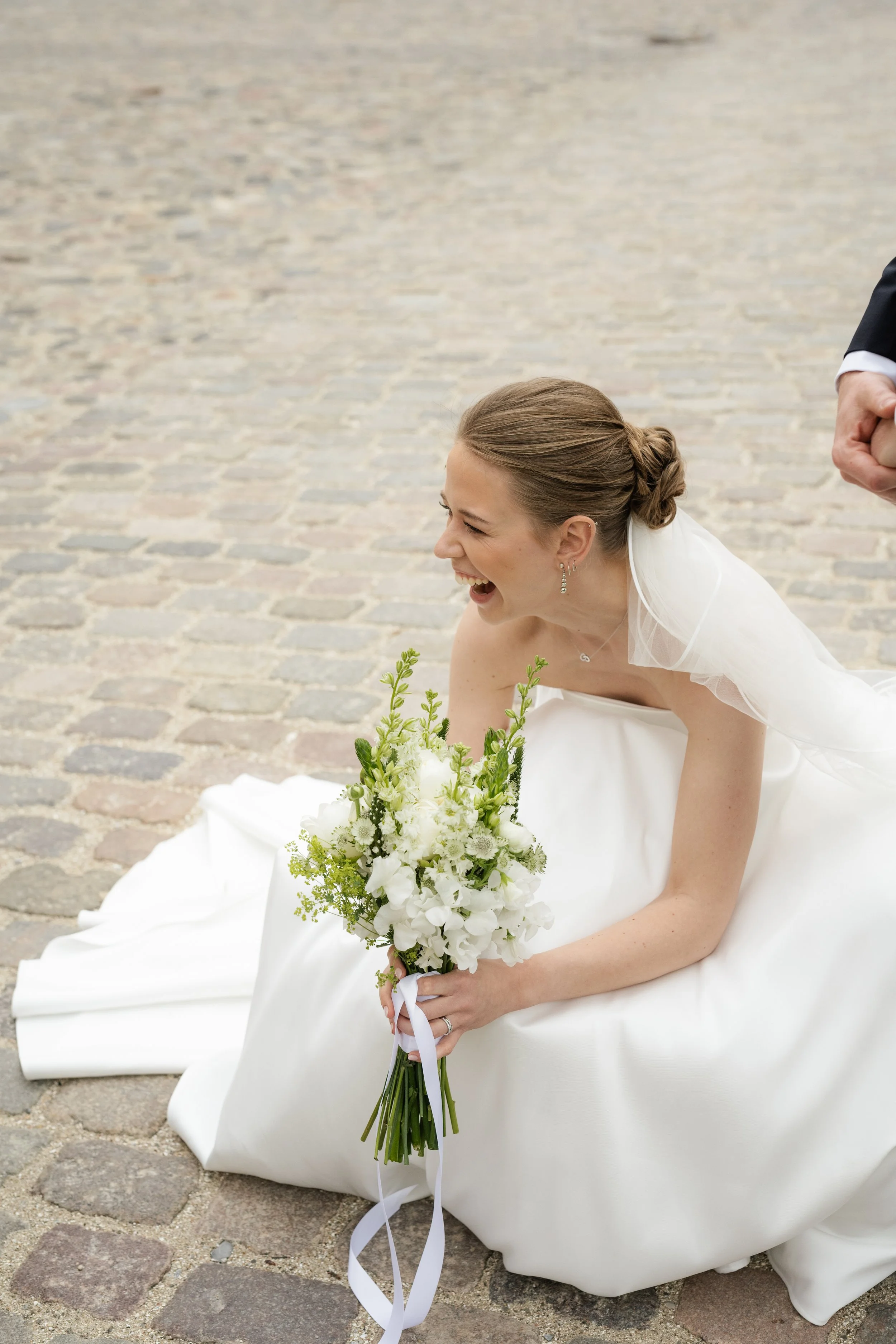 A smiling bride in a white wedding dress holding a bouquet of white flowers, sitting on a cobblestone street.