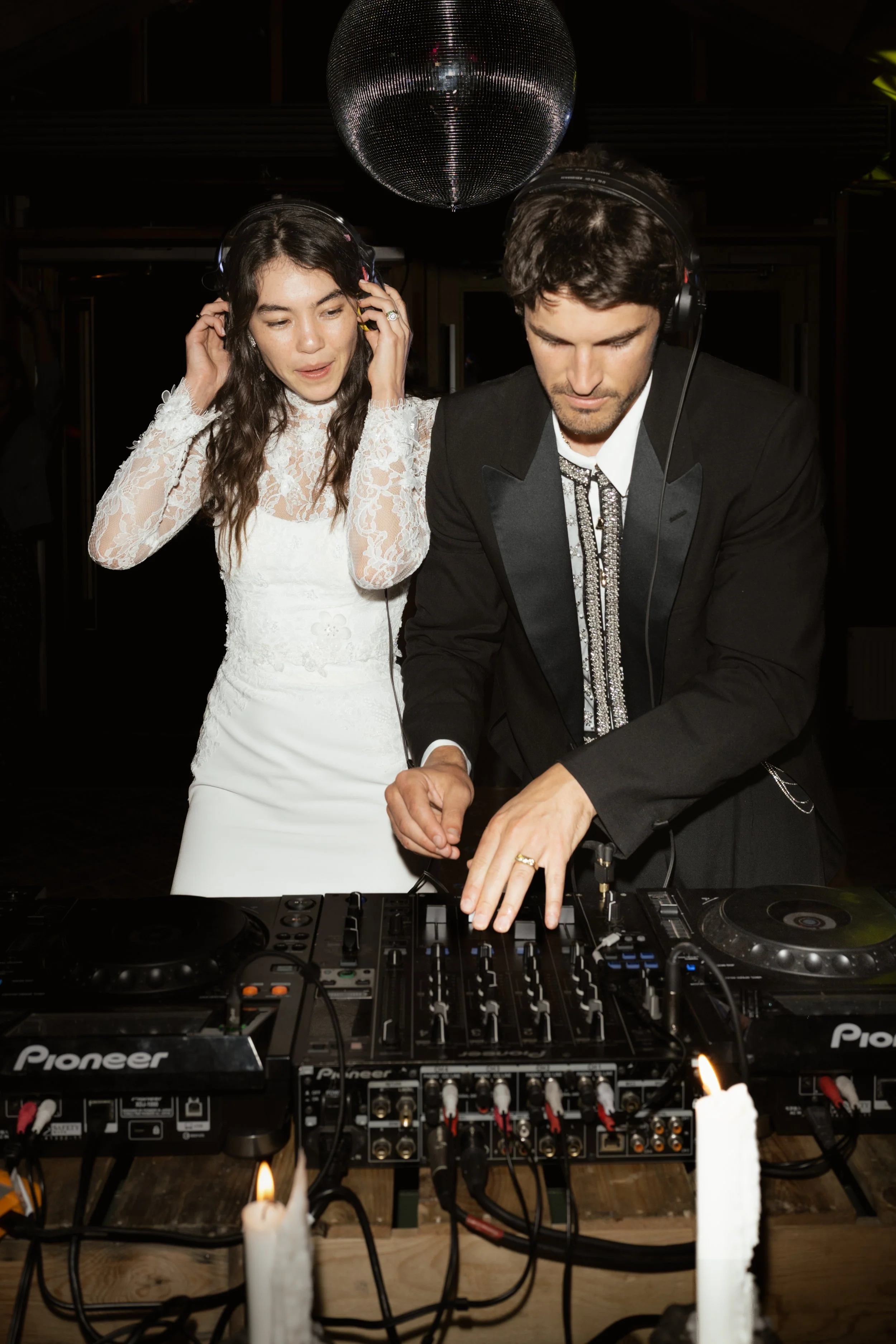 A bride and groom at a wedding reception, with the groom DJing with professional DJ equipment and the bride listening to music with headphones, under a disco ball.