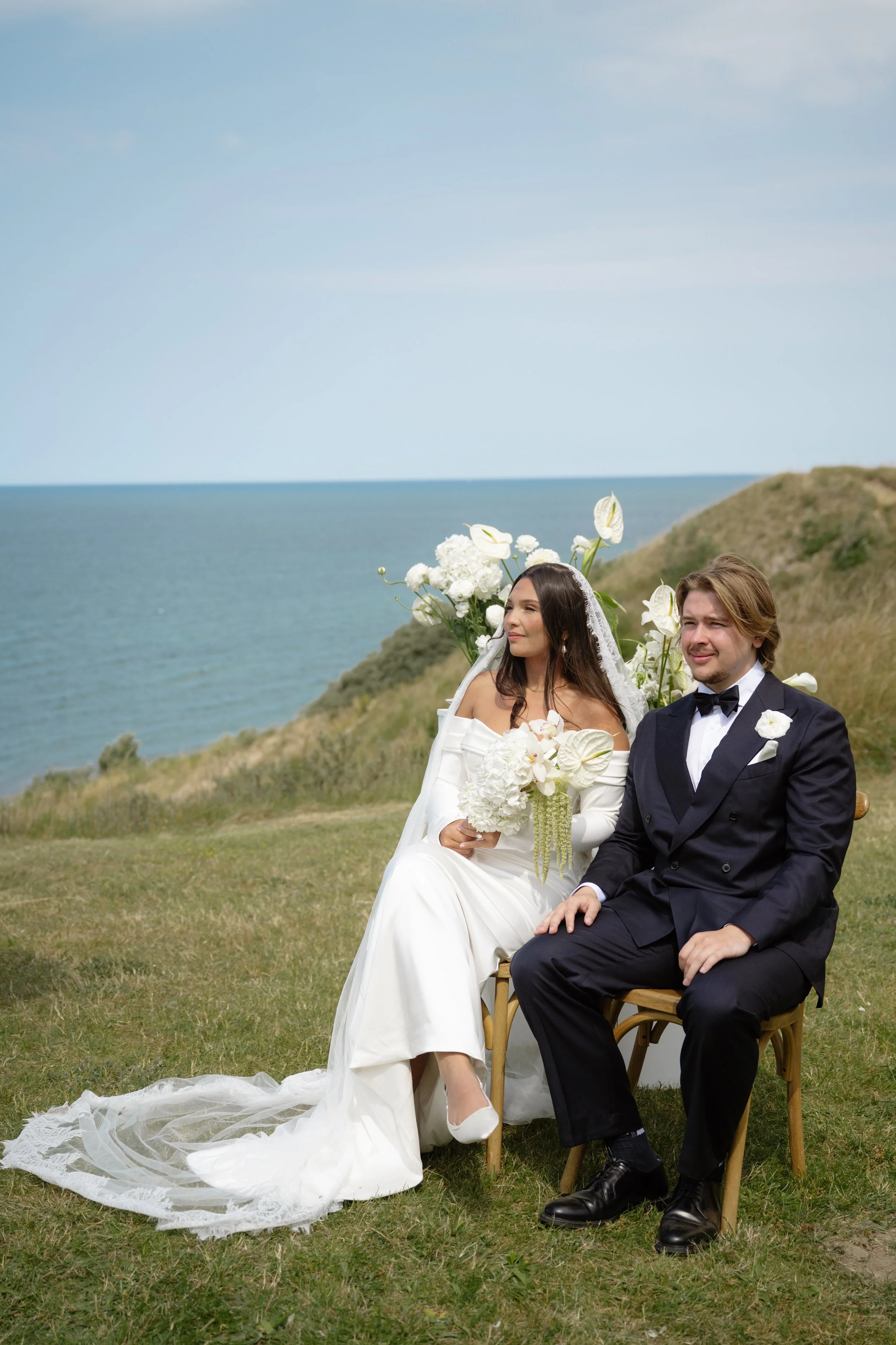 A bride and groom sit on chairs outdoors near the ocean during a wedding ceremony with a backdrop of grassy hills and blue sky.