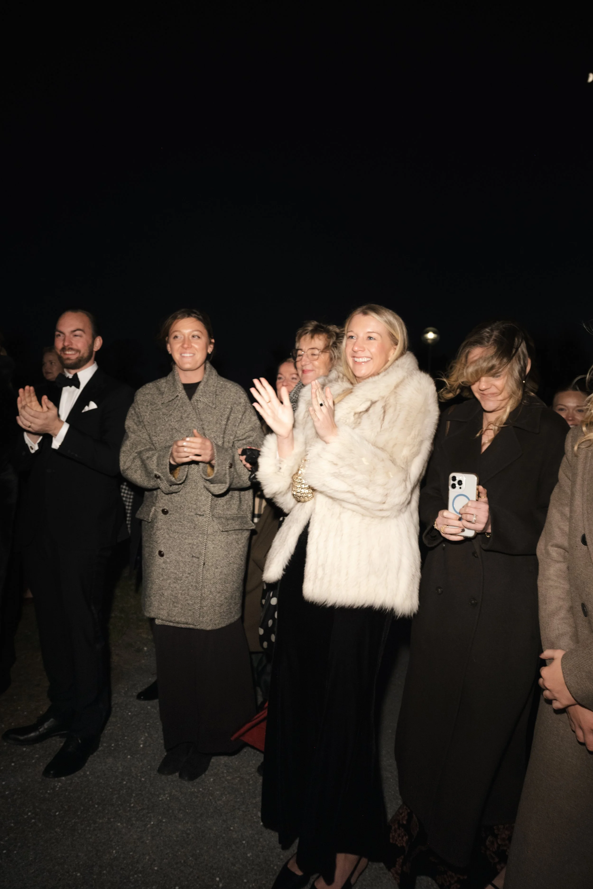 People standing outdoors at night, dressed in formal clothing, some smiling and clapping, with some using phones to take pictures.