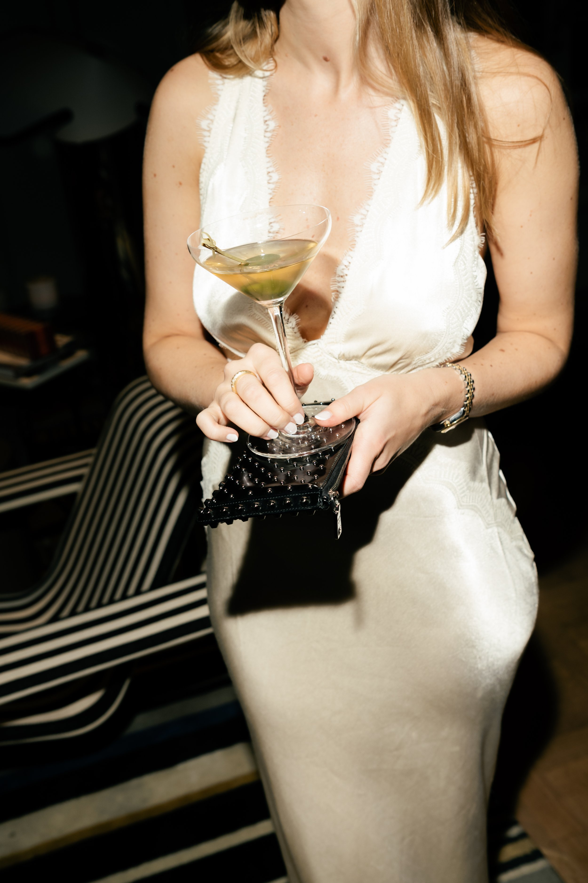 A woman in a cream-colored dress holding a martini glass with a green olive, standing indoors with striped furniture in the background.