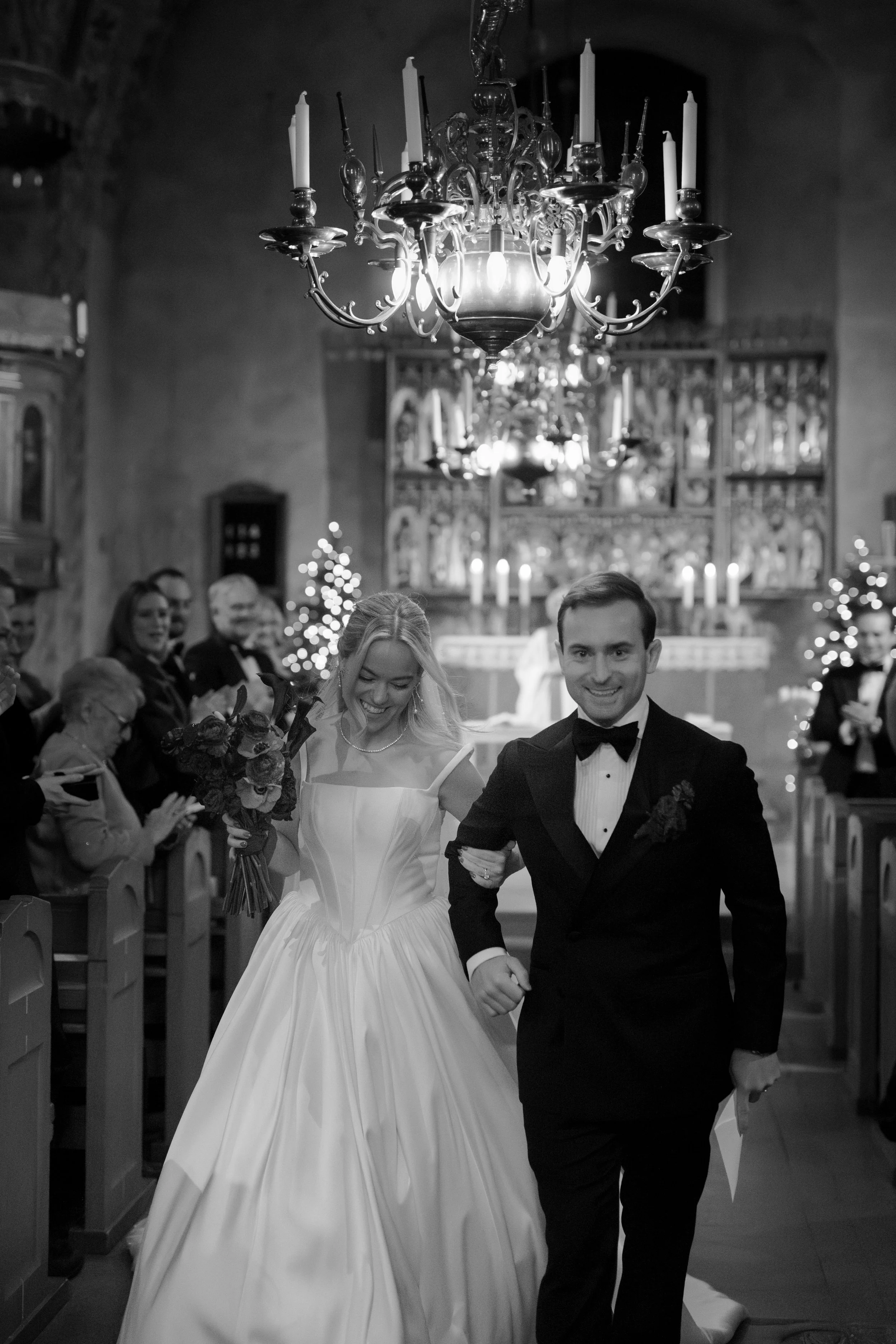 A black and white photo of a bride and groom walking down the aisle inside a church, with guests clapping and smiling in the background, Christmas trees with lights, and a grand chandelier above.