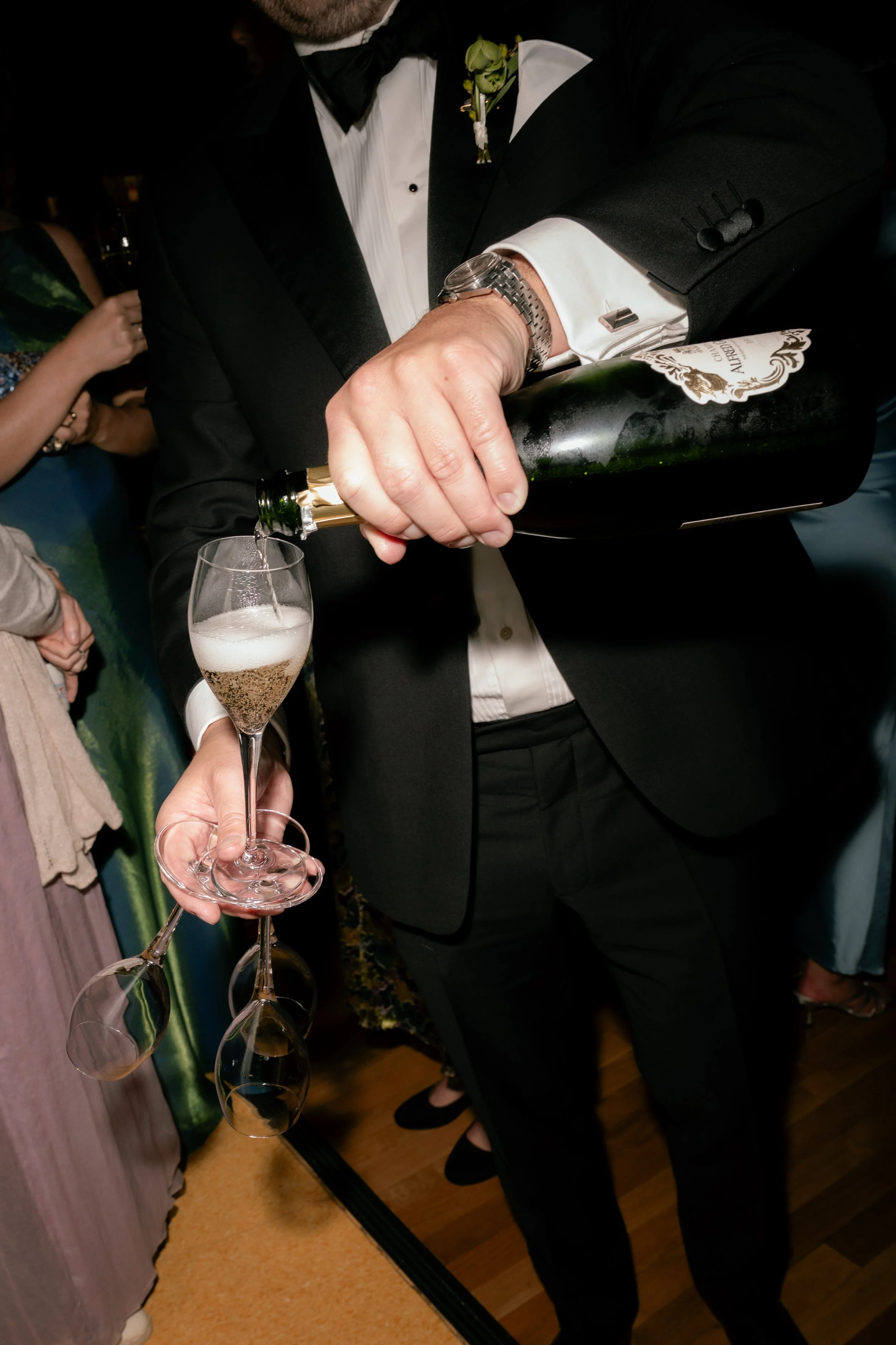 A man in a black tuxedo, white shirt, and black bow tie is pouring champagne into a flute glass, holding a bottle with a white and gold label. In the background, other guests are visible at an indoor event.