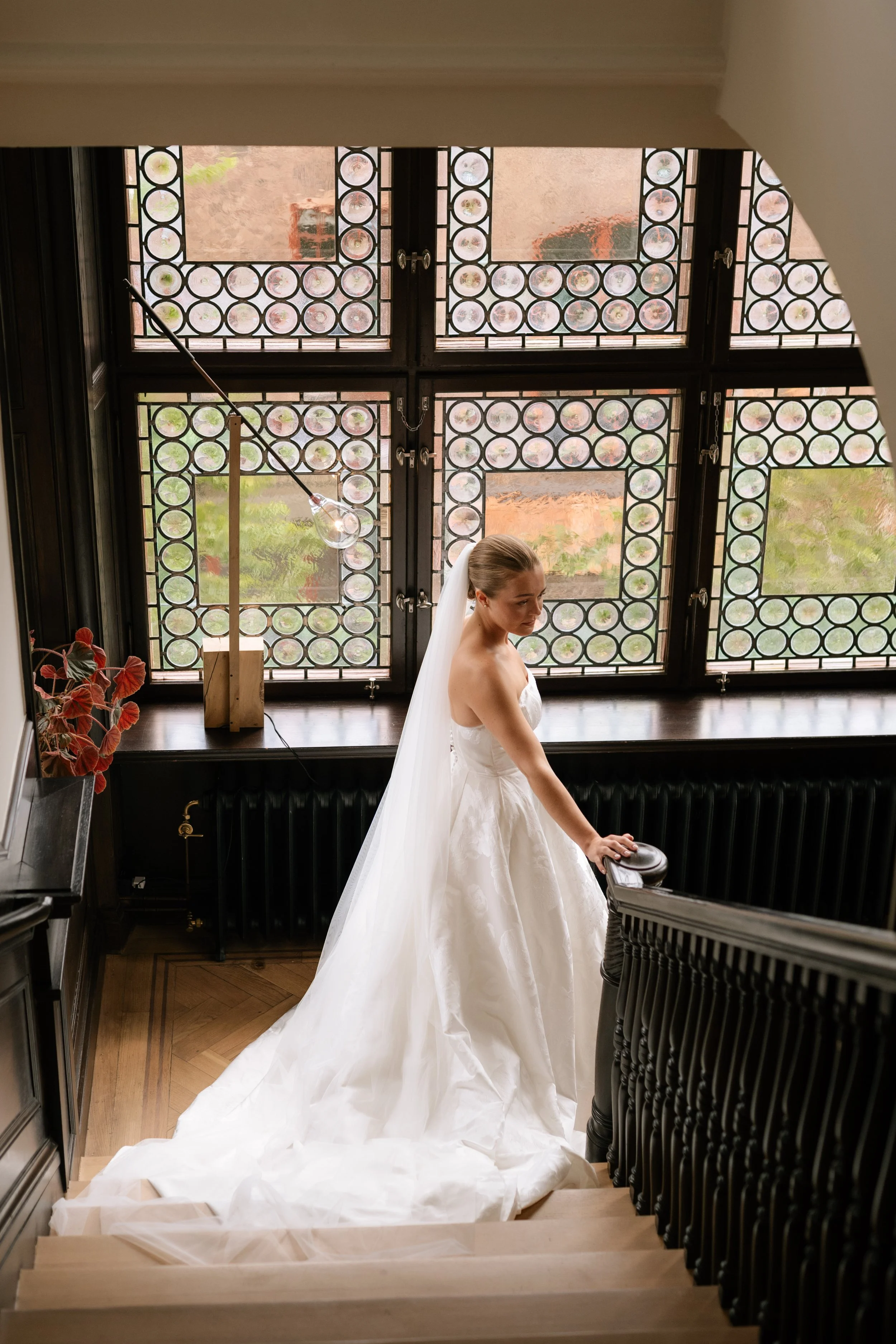 A bride in a white wedding gown and veil standing on a staircase inside a building, holding onto the railing, with decorative windows in the background.