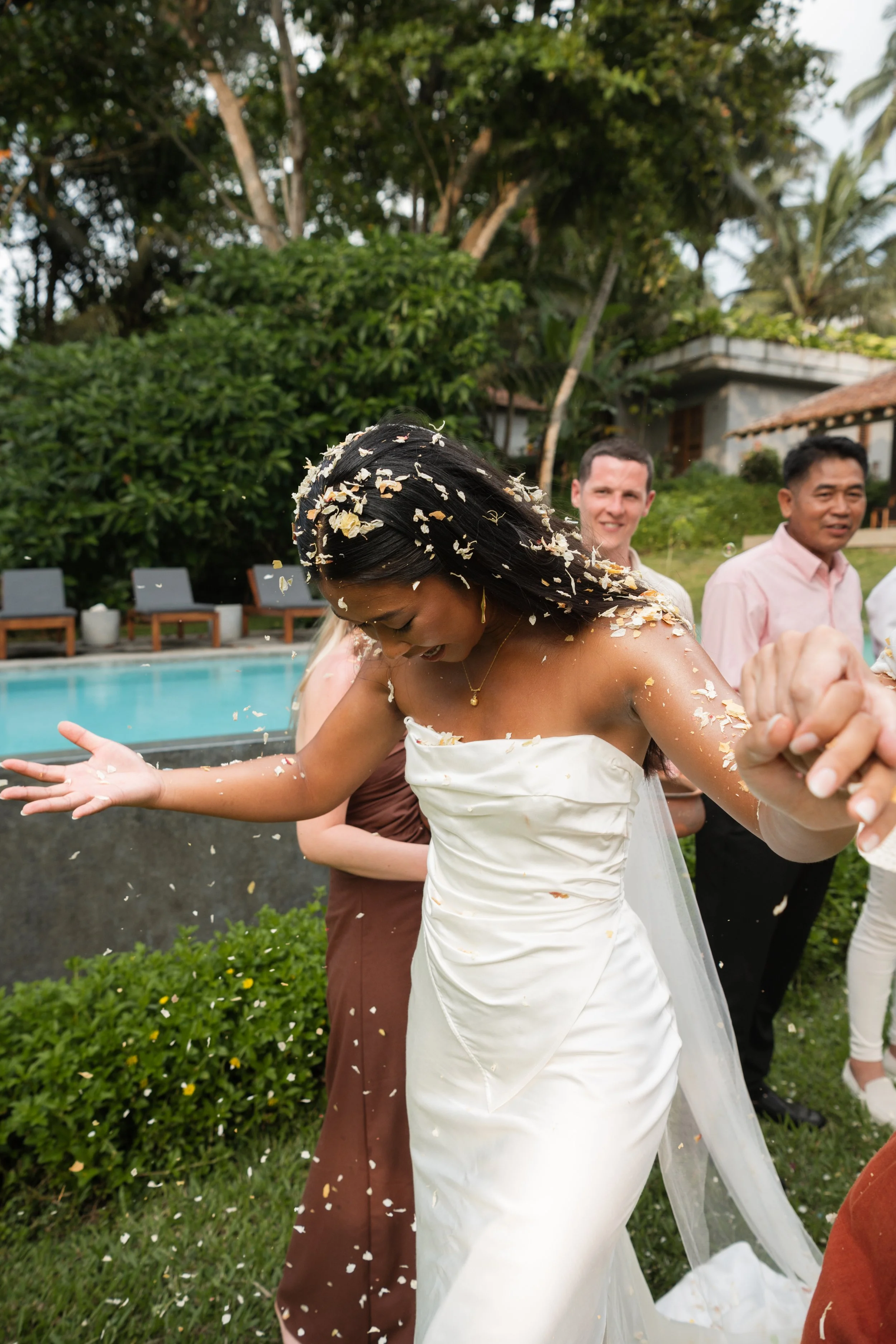 Bride in a white wedding dress celebrating outdoors with guests, surrounded by greenery, as flower petals are showered over her.