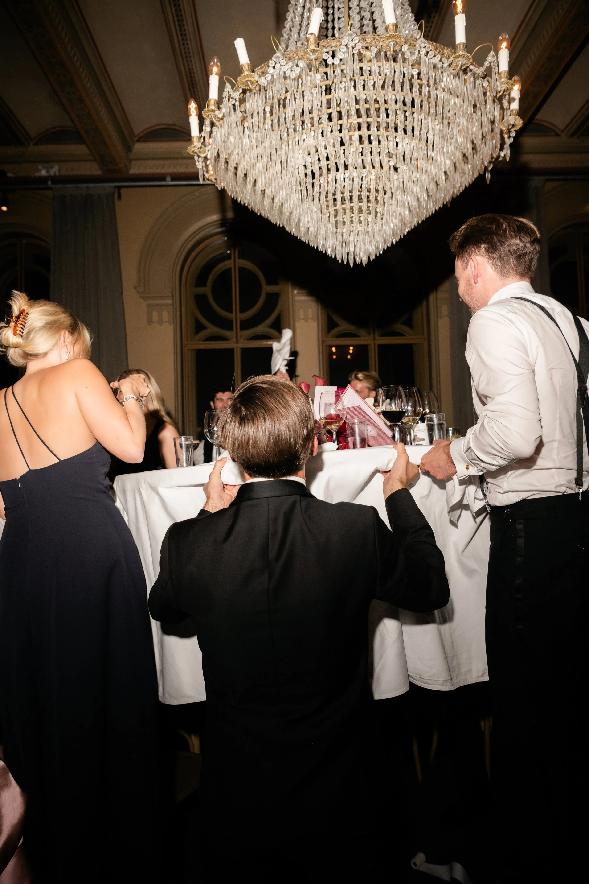 People at a formal event, with a woman in a black dress, a man in a tuxedo, and another man in a white shirt, under a large crystal chandelier, with a table set with wine glasses and a pink gift bag.