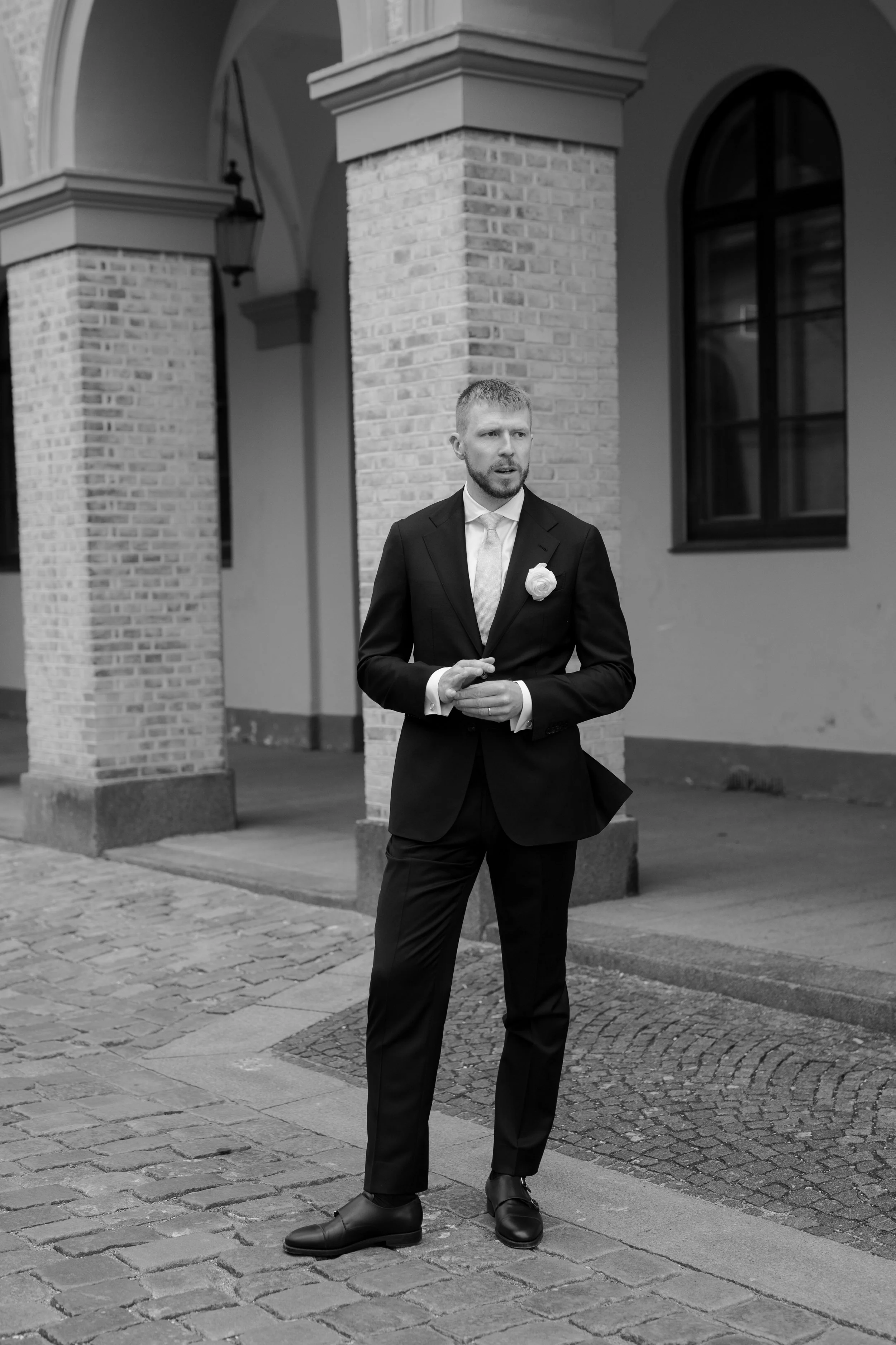 A man in a tuxedo with a boutonniere stands outdoors on a cobblestone sidewalk in front of brick pillars and arched windows, possibly preparing for a formal event or wedding.