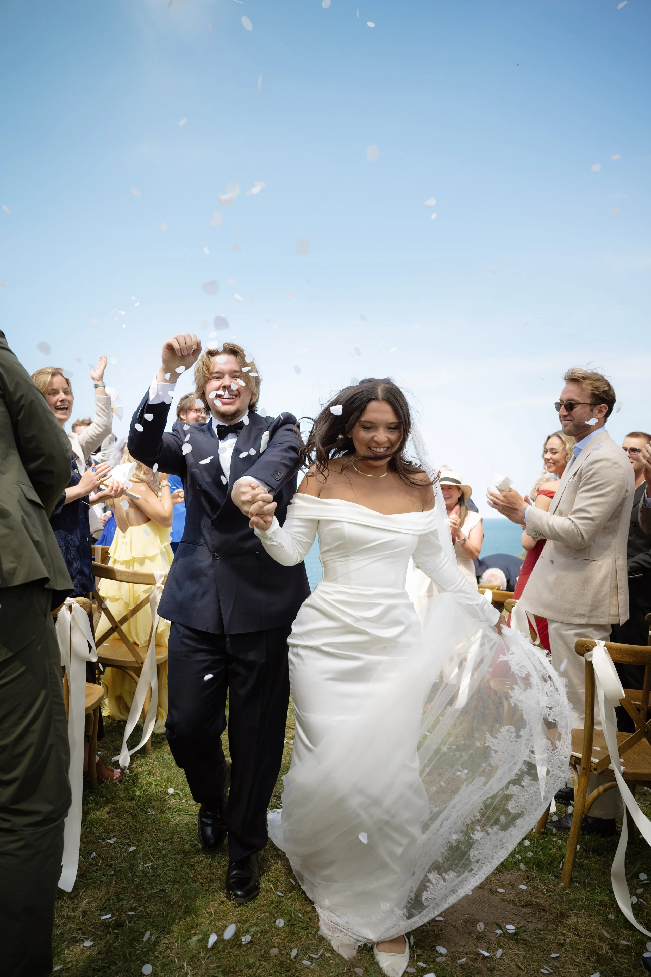 A newlywed couple walking down the aisle at their outdoor wedding, celebrating with guests throwing confetti, against a bright sky with ocean in the background.