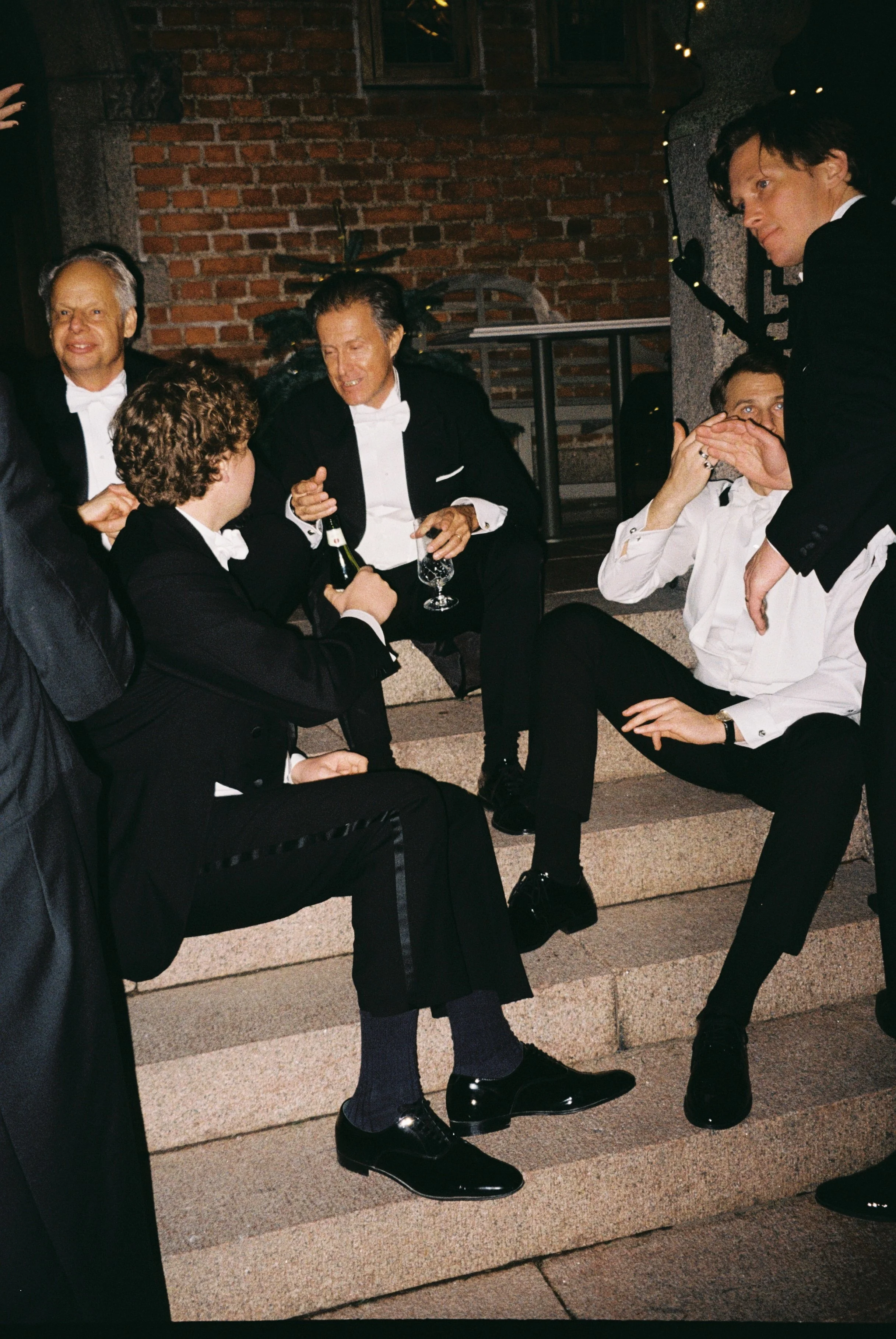 A group of five men in tuxedos sitting on outdoor steps, engaging in conversation, with some holding drinks, at a formal event during the evening.